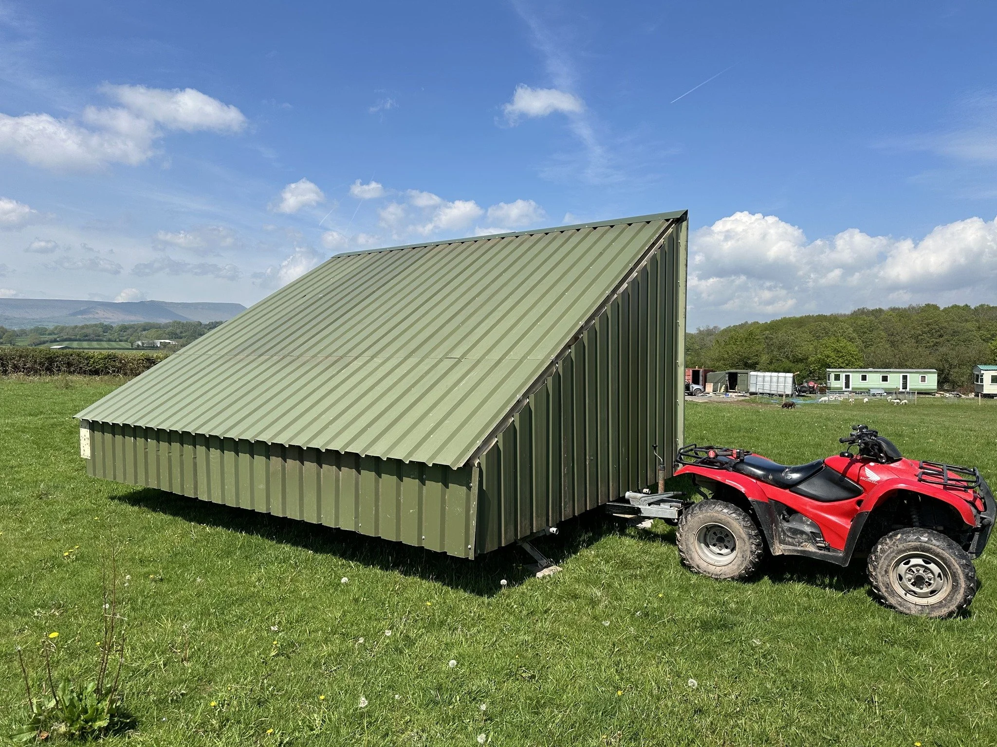 A green metal shed with a sloped roof, situated on a grassy field under a partly cloudy sky, with a red ATV parked beside it.