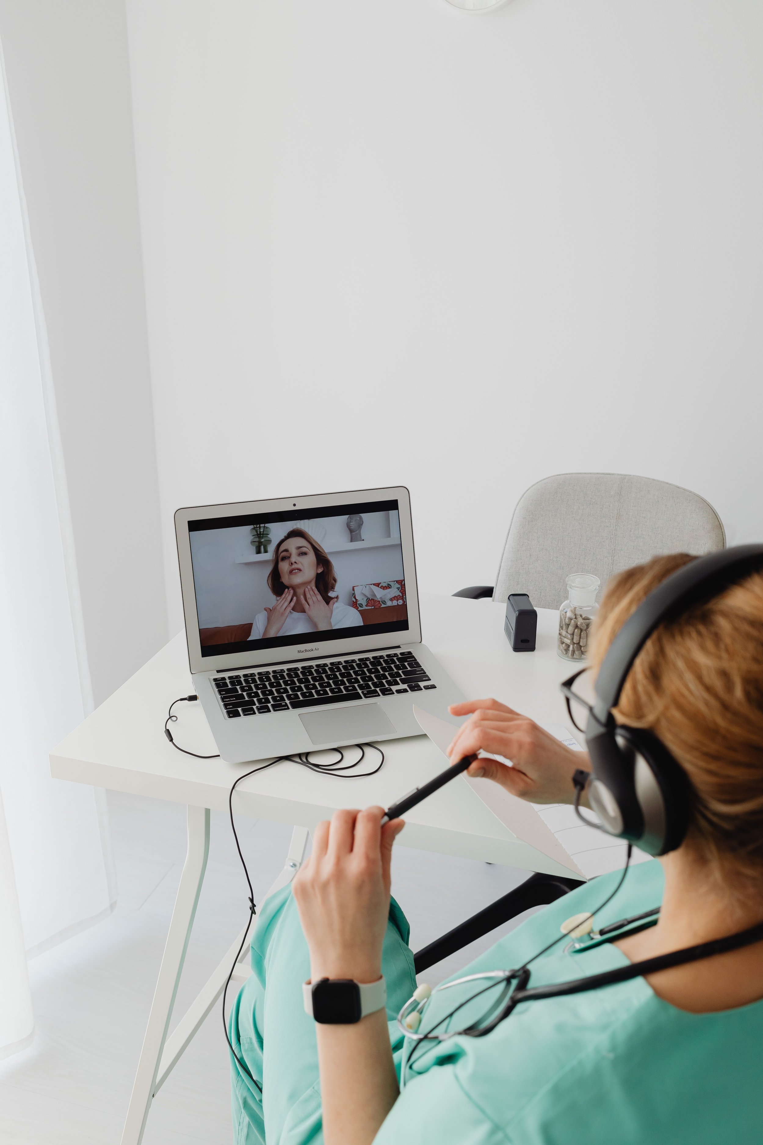A healthcare professional in scrubs and a stethoscope wearing headphones, participating in a video call with a woman on a laptop in a clean, white, minimalistic room.