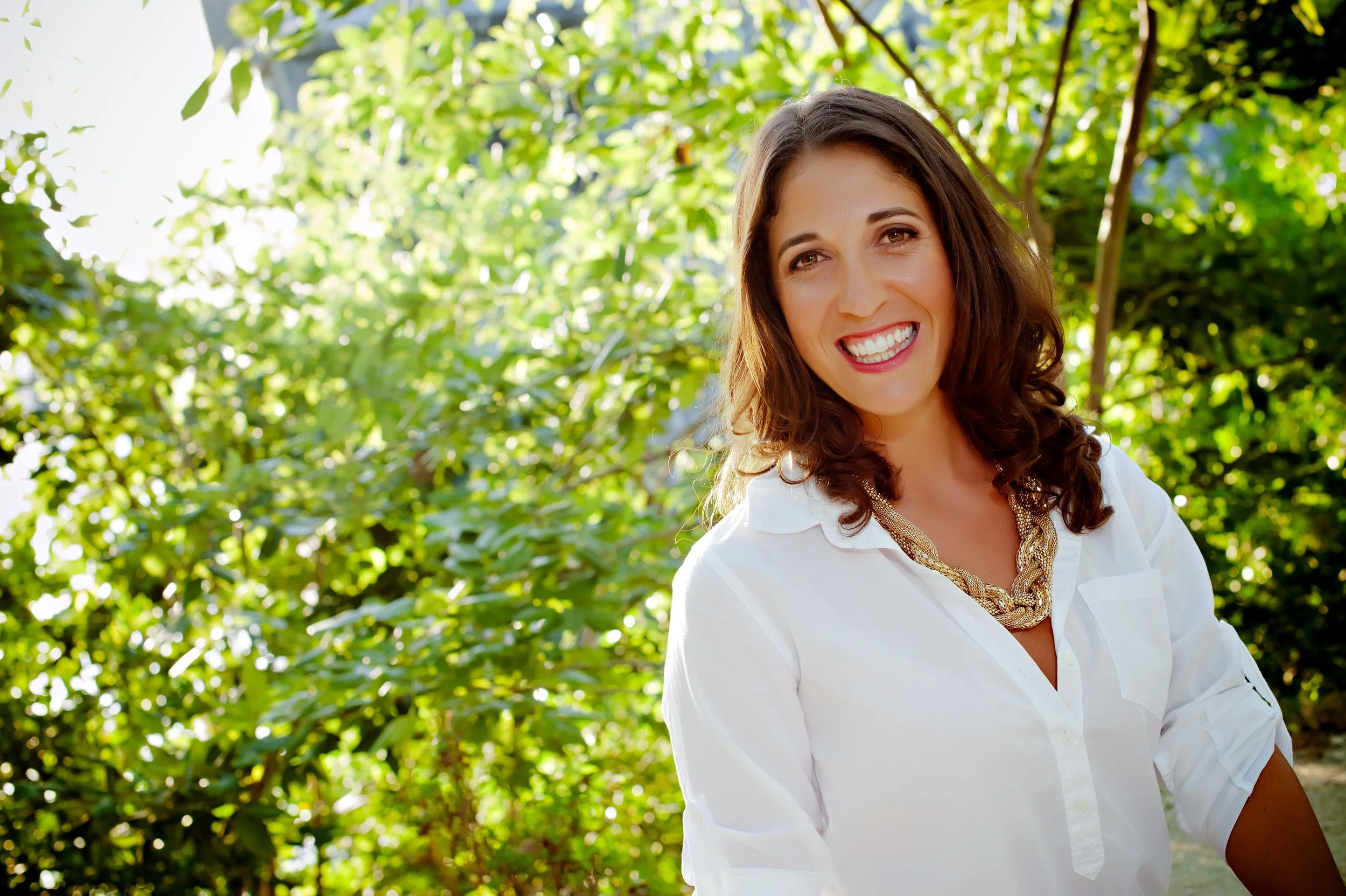 A woman with long brown hair and a big smile, wearing a white shirt and a gold necklace, standing outdoors with green leafy trees in the background.