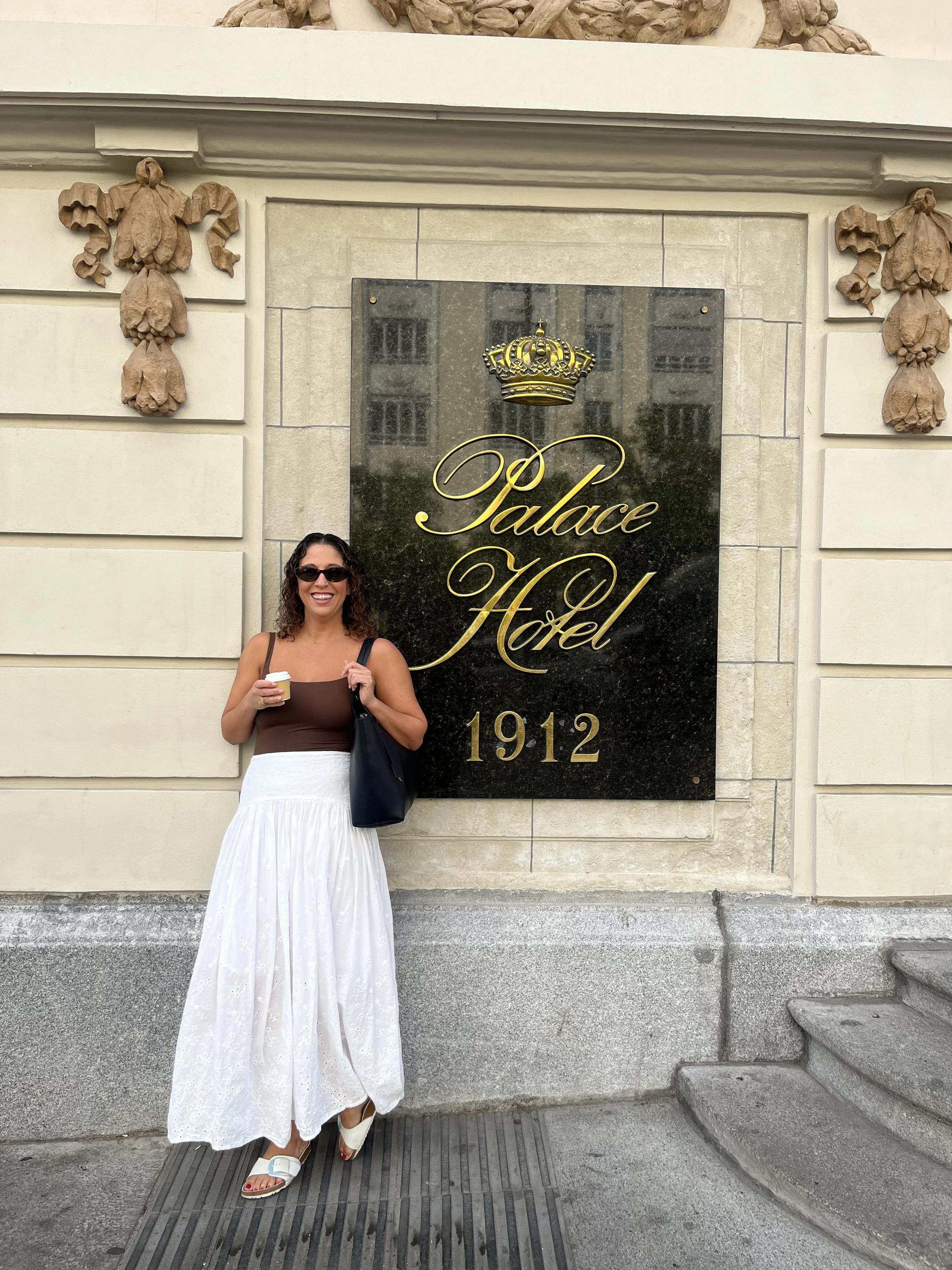 A woman with curly hair wearing sunglasses, a brown sleeveless top, and a long white skirt with eyelet details, standing in front of a building. She is holding a cup of coffee in her right hand and a black bag over her left shoulder, smiling at the camera. The background features a black sign with gold lettering that reads 'Palace Hotel' and a crown emblem, with the year '1912' below. The building has beige stone walls with decorative stone floral elements and a stone staircase to her right.