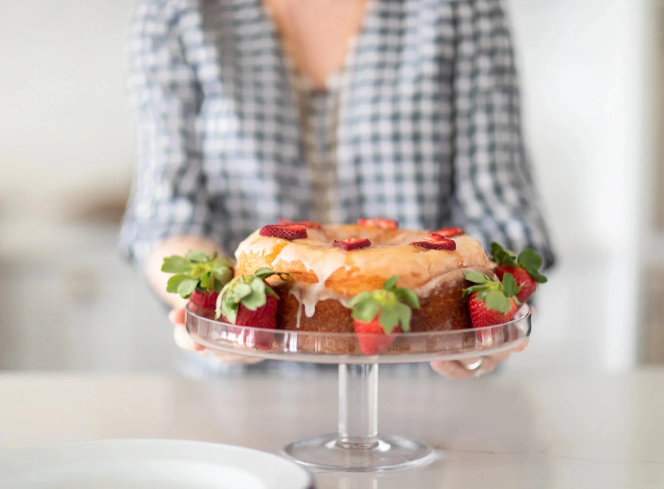 A person in a checkered shirt holds a glass cake stand with a layered dessert topped with strawberries, garnished with green herbs.