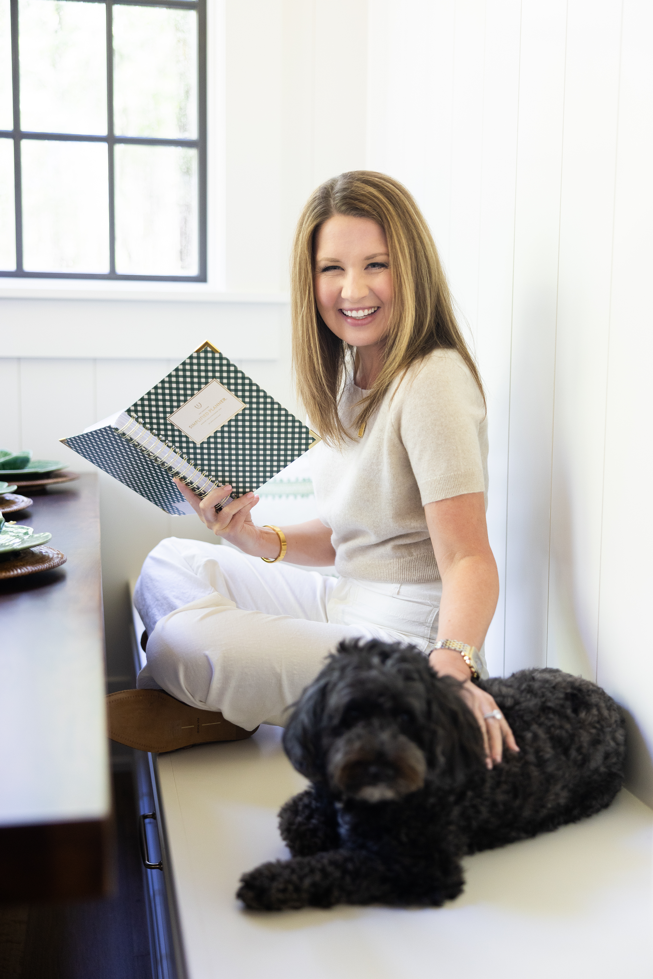 A woman with brown hair wearing a beige top and white pants is sitting cross-legged on a white floor, holding a black and white checkered notebook, and smiling while looking to her right. She is in a bright room with white walls and a window. A large, black, curly-haired dog is lying at her feet, partially visible.