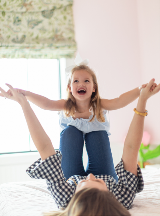 A young girl with a big white bow in her hair, laughing and playing with an adult lying on a bed indoors, with sunlight coming through a window.