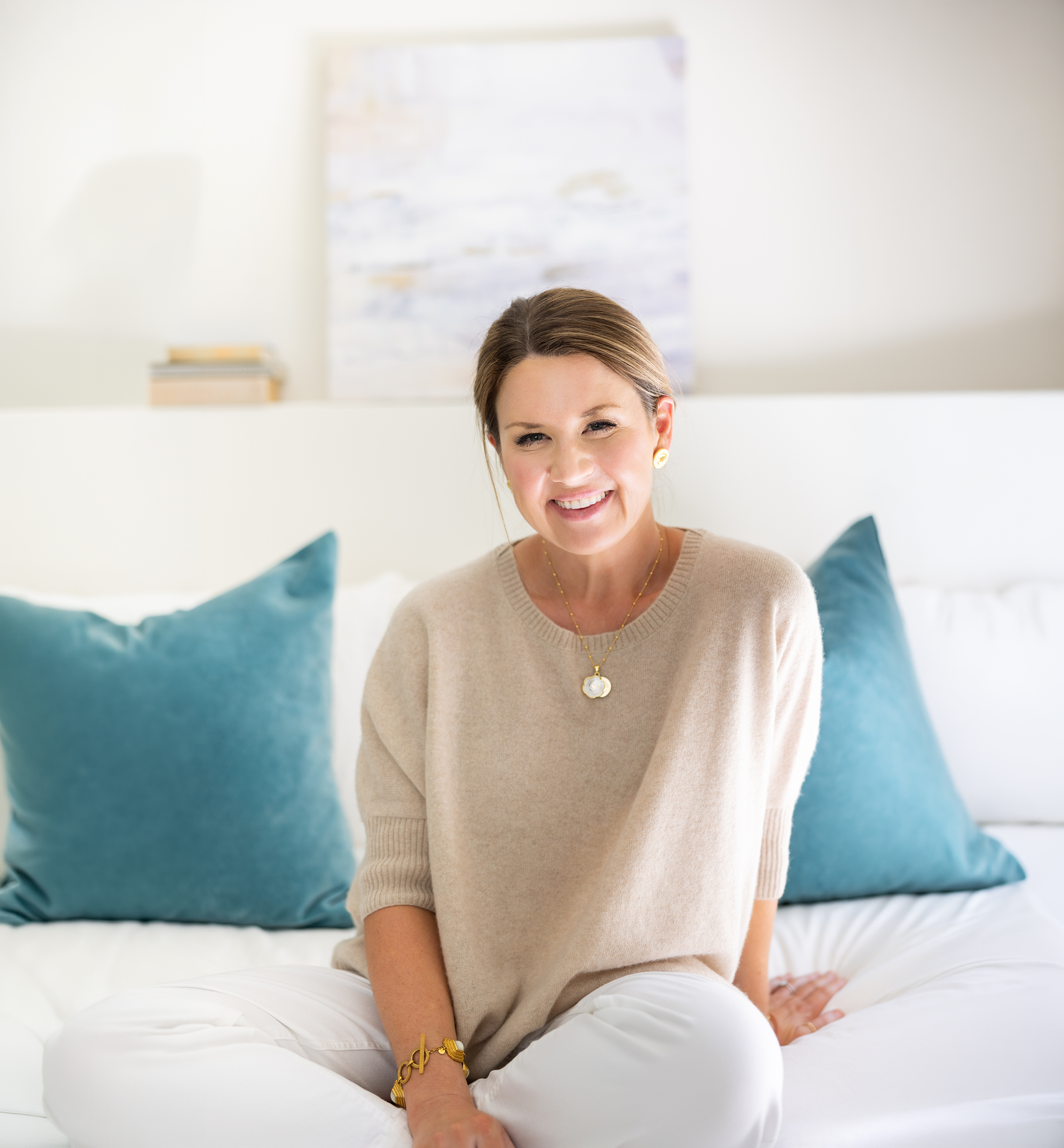 A woman with light brown hair tied back, smiling and sitting on a bed with white sheets and blue cushions. She is wearing a beige sweater, white pants, and gold jewelry, including a pendant necklace, earrings, and a bracelet.