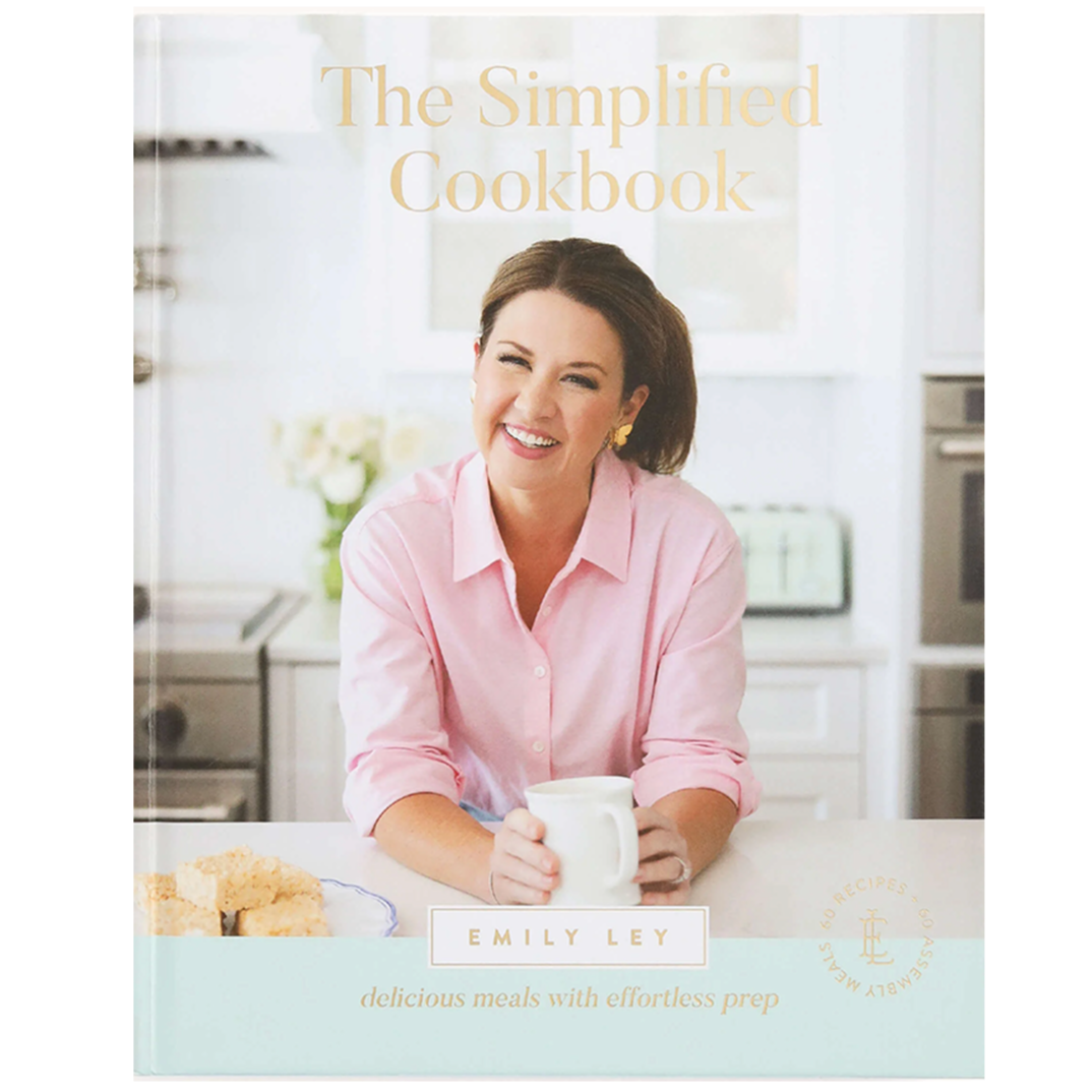 Cover of The Simplified Cookbook by Emily Ley featuring a woman in a pink shirt smiling and holding a mug in a bright kitchen, with baked goods on the counter.