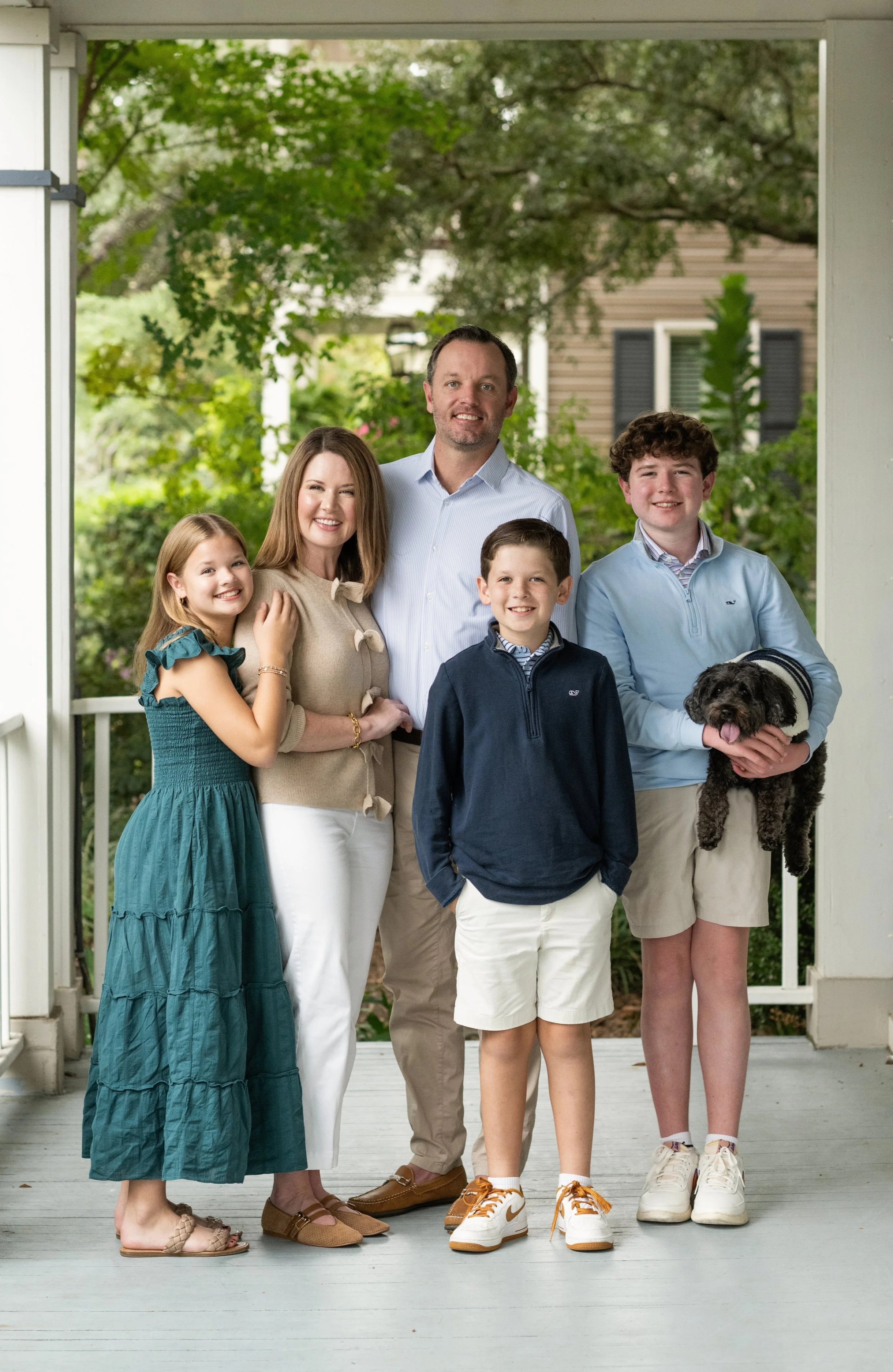 A family of six people and a dog standing on a porch with greenery in the background. They are smiling and dressed casually.