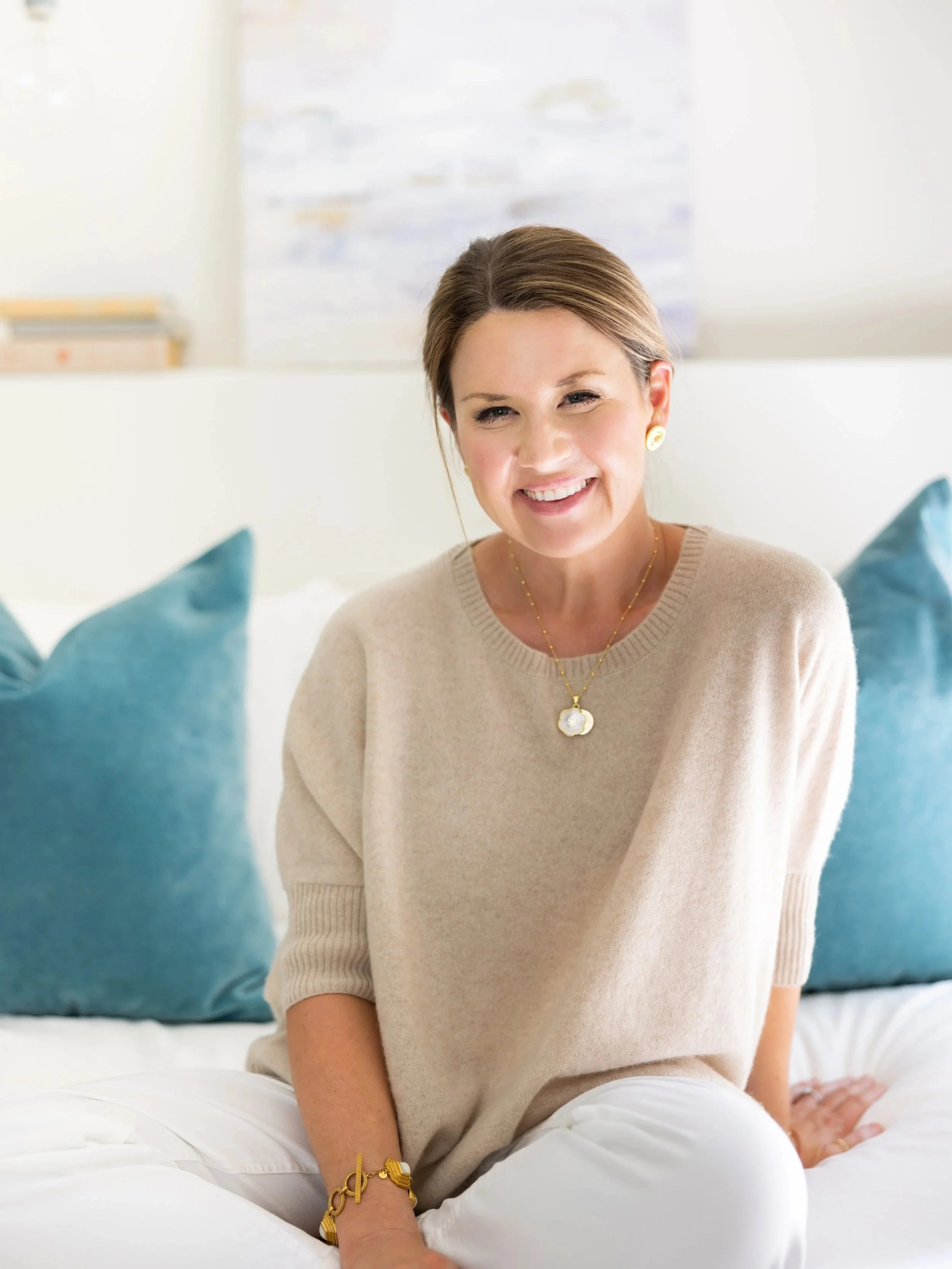 A woman sitting on a bed with pillows, smiling at the camera, wearing a beige sweater and gold jewelry in a bright room.