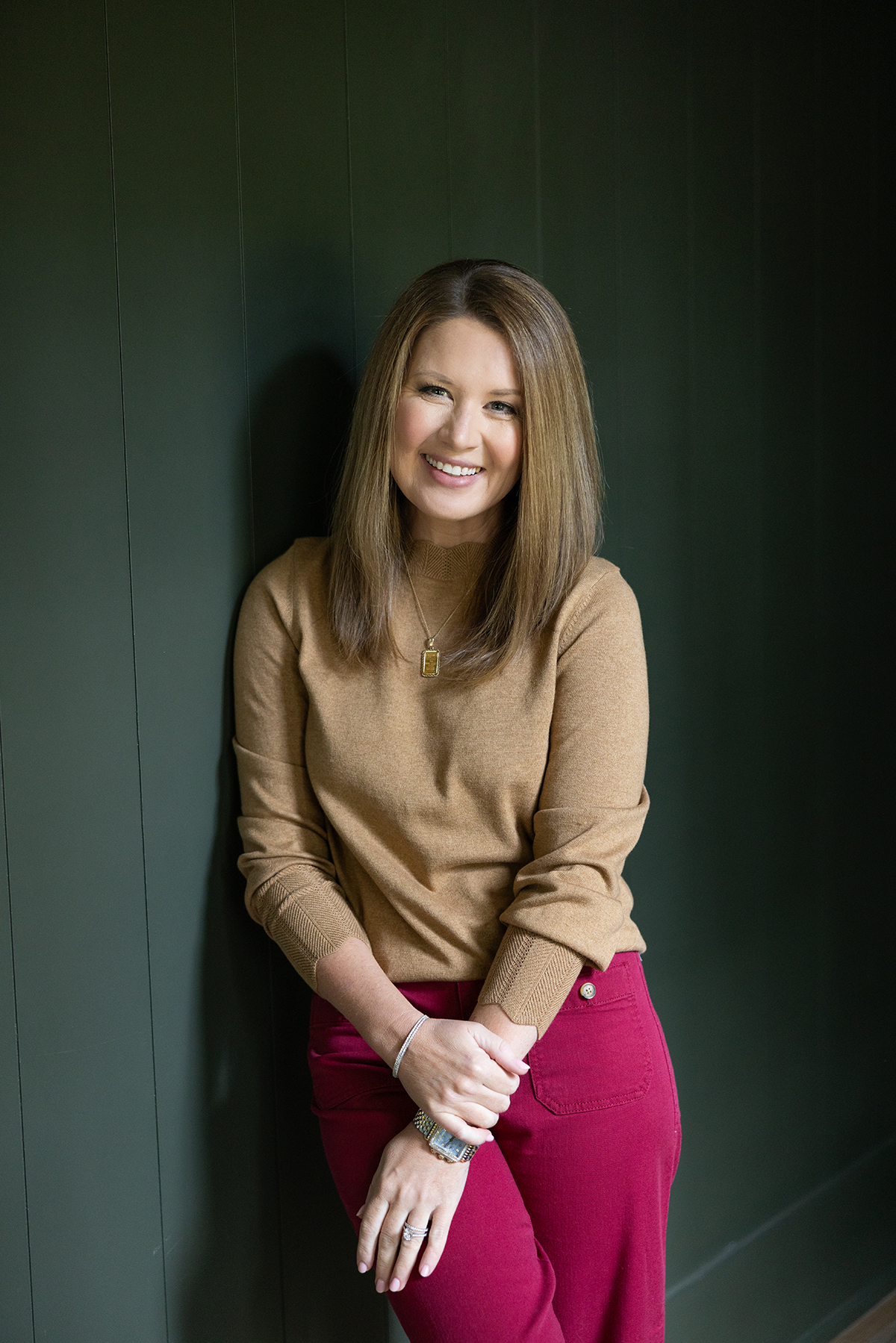 A woman with long light brown hair smiling and holding a patterned notebook, sitting on a white bench with a large black and brown dog lying at her feet in a bright room with white walls and a window in the background.