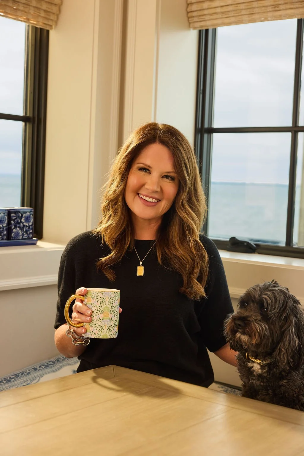A woman with long wavy brown hair smiling and holding a colorful mug, sitting at a table with a black dog beside her, in a room with large windows overlooking water.
