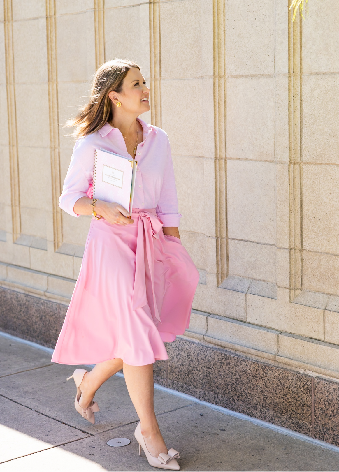 A woman wearing a pink dress, pink cardigan, and high heels, holding a notebook and walking on a city sidewalk.