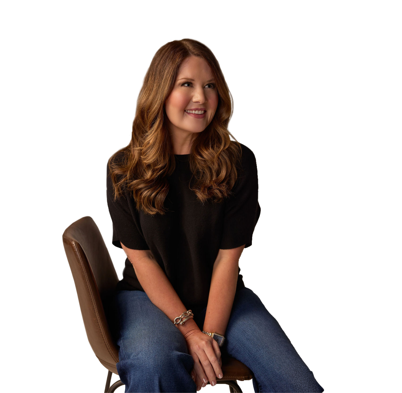 A woman with long wavy brown hair sitting on a brown chair, smiling, wearing a black top and blue jeans.