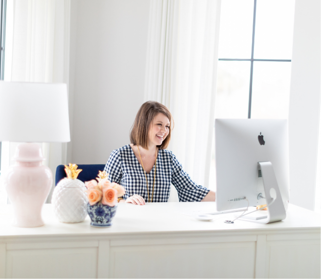 Woman with shoulder-length brown hair smiling while working at a desk in a bright room with white walls and large windows, surrounded by decorative vases and a lamp.