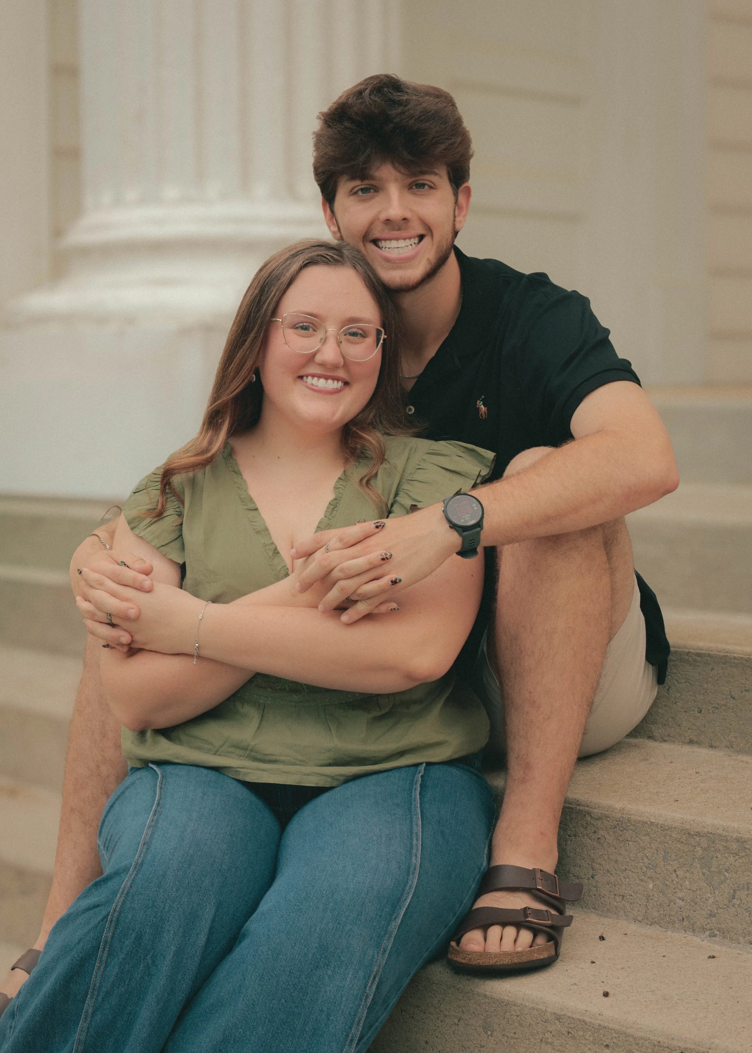 A young man and woman sitting on steps, smiling, with the man hugging the woman from behind.