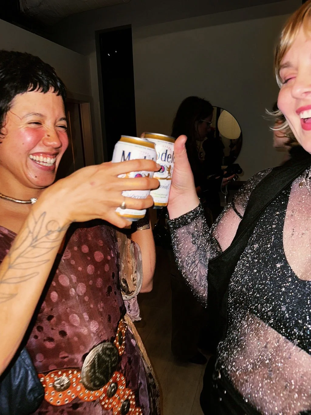 Two women laughing and toasting with beer cans at a party, one with short black hair and tattooed arm, the other with blond hair wearing a glittery top.
