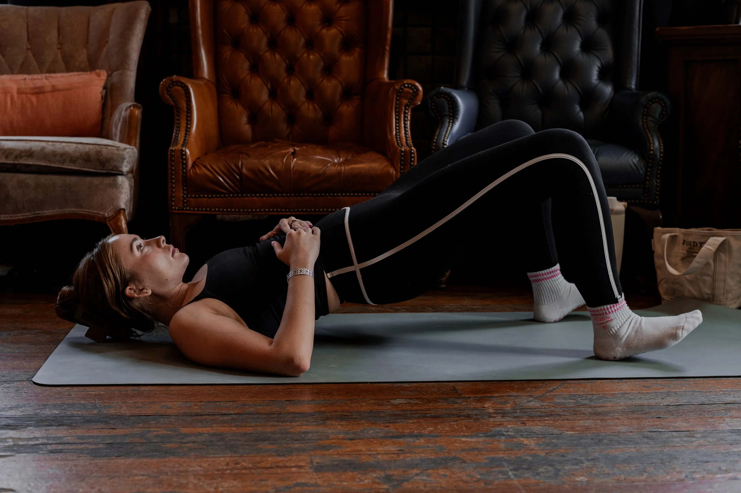 A woman with red hair lies on her back on a yoga mat, holding her knees to her chest, in a dimly lit room with vintage leather furniture and wooden flooring.