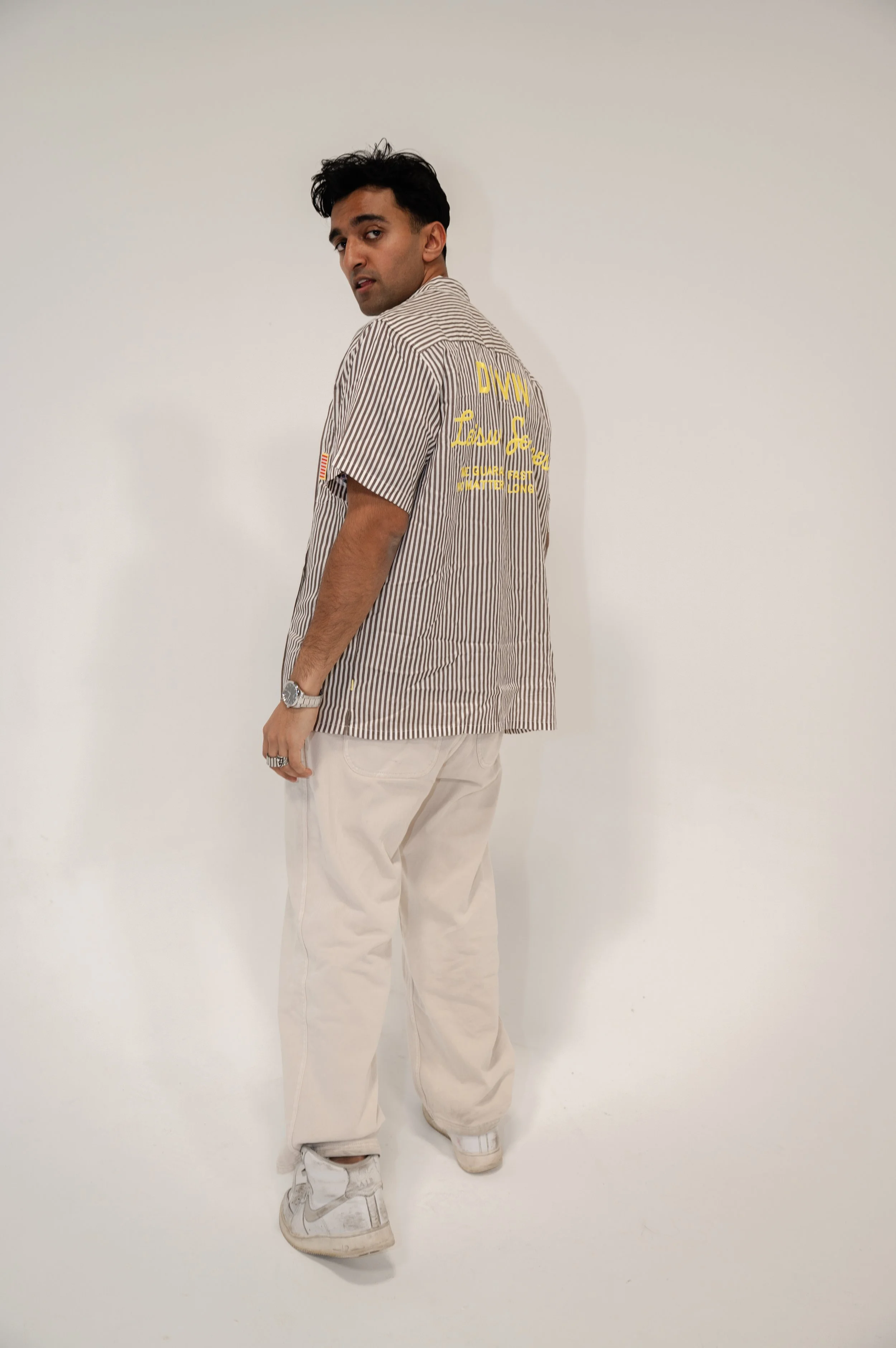 A man with dark hair and a wristwatch, wearing a striped shirt with yellow text on the back and white pants, stands against a plain white background, looking over his shoulder.
