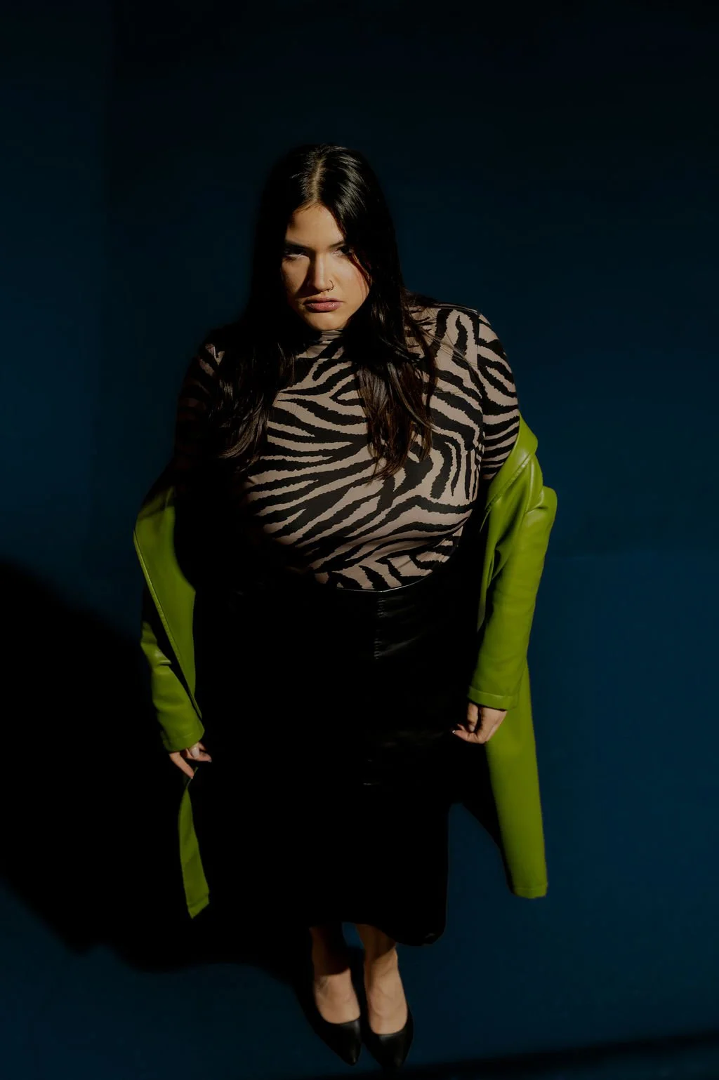 A woman with long dark hair wearing a zebra print top, a black skirt, black heels, and a green coat draped over her shoulders standing against a dark background.