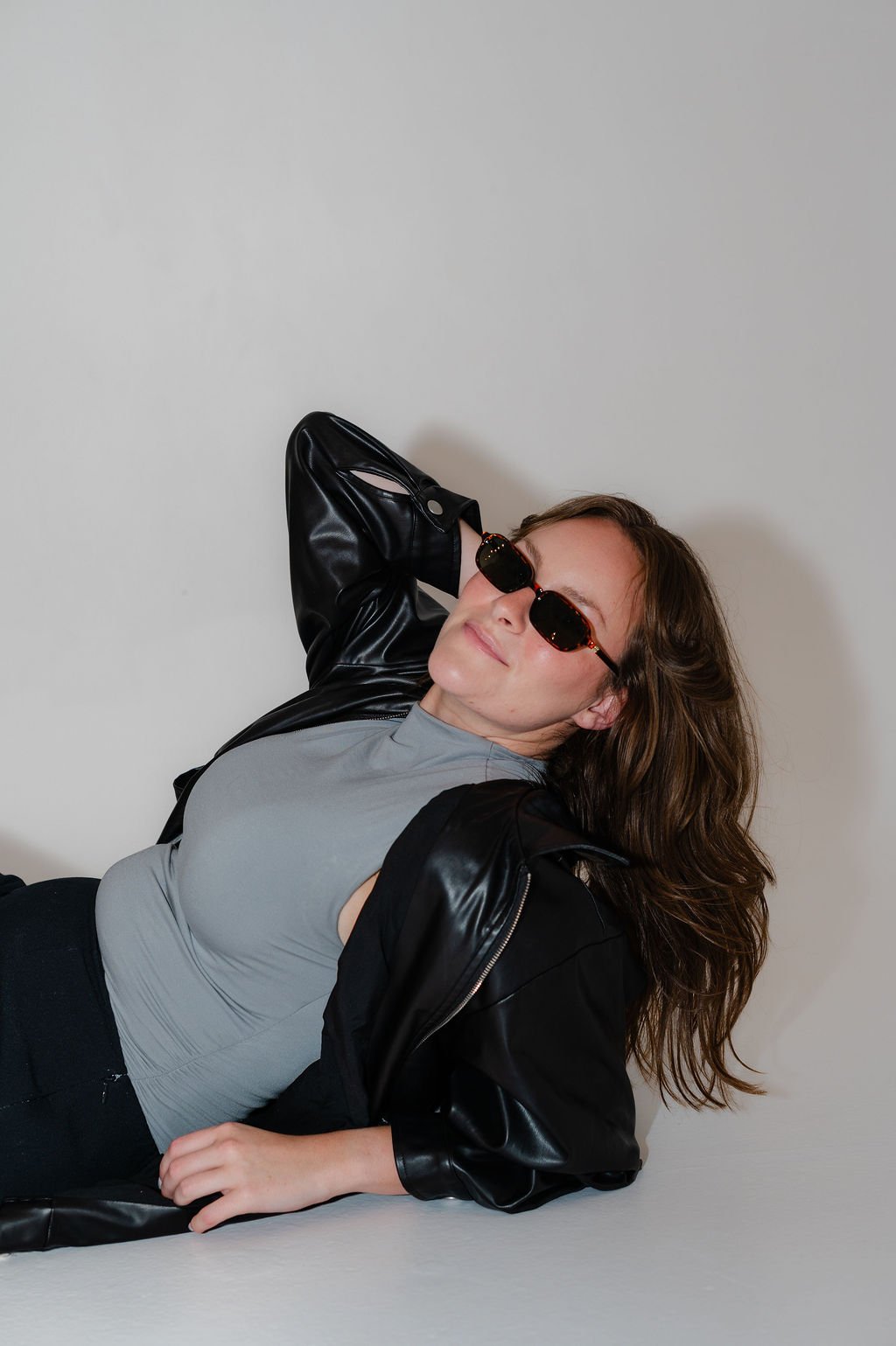 A woman with wavy brown hair wearing black sunglasses, a gray T-shirt, and a black leather jacket, relaxing against a plain white wall.