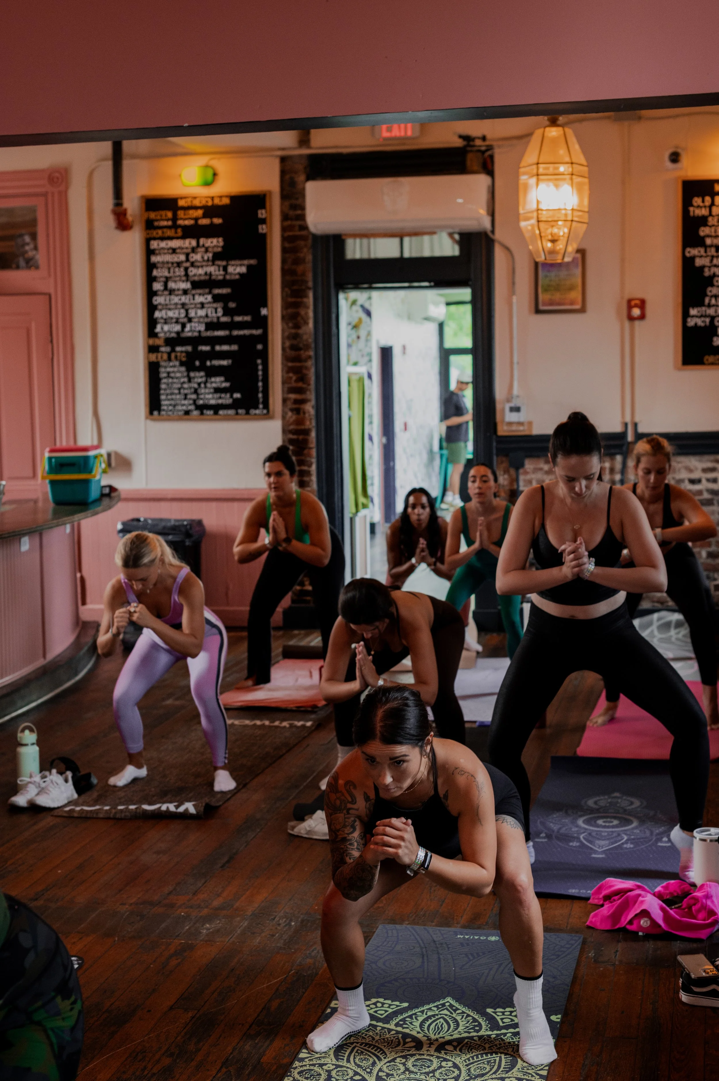 Group of people practicing yoga indoors, performing a prayer pose, with some in downward dog position, on colorful mats. The room has wooden floors, decorative walls, and soft lighting.