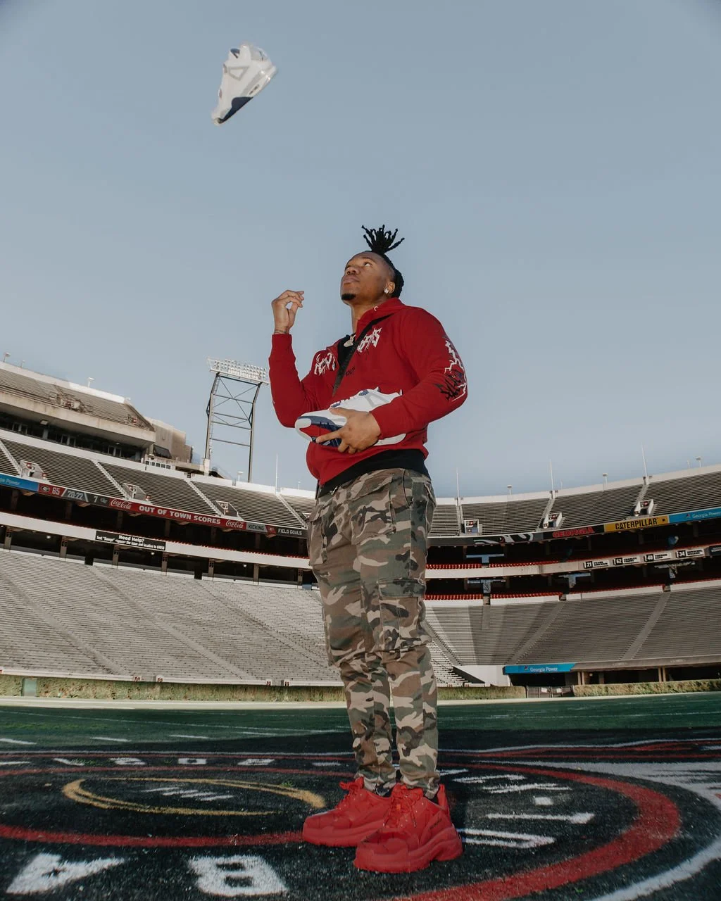 A man with dreadlocks tossing a white sneaker into the air at a football stadium, wearing a red hoodie, camouflage pants, and red sneakers.