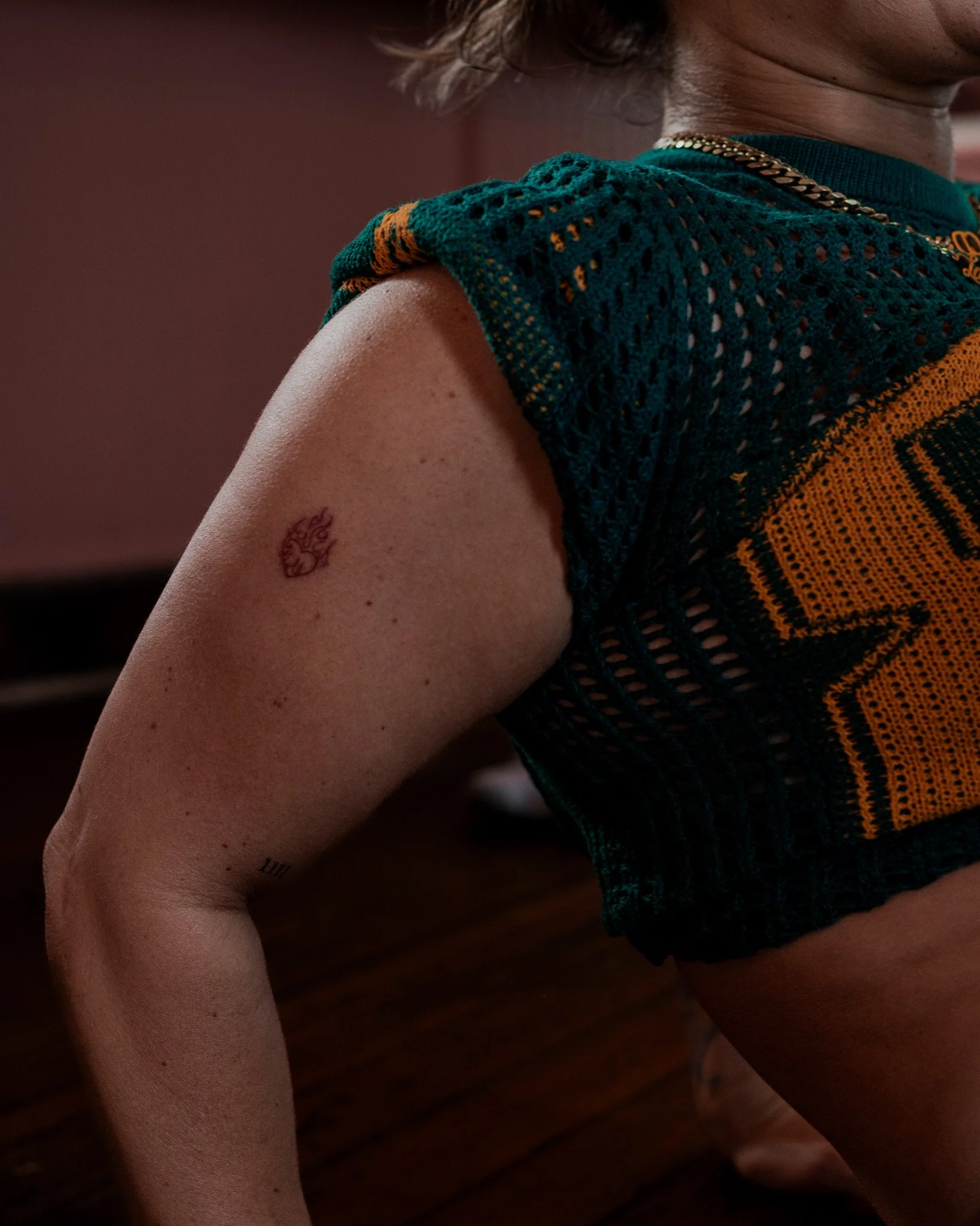 Close-up of a person's shoulder, tattooed arm, and part of their face, wearing a teal and orange knitted sleeveless shirt and a gold chain necklace. The background is blurred and dark.