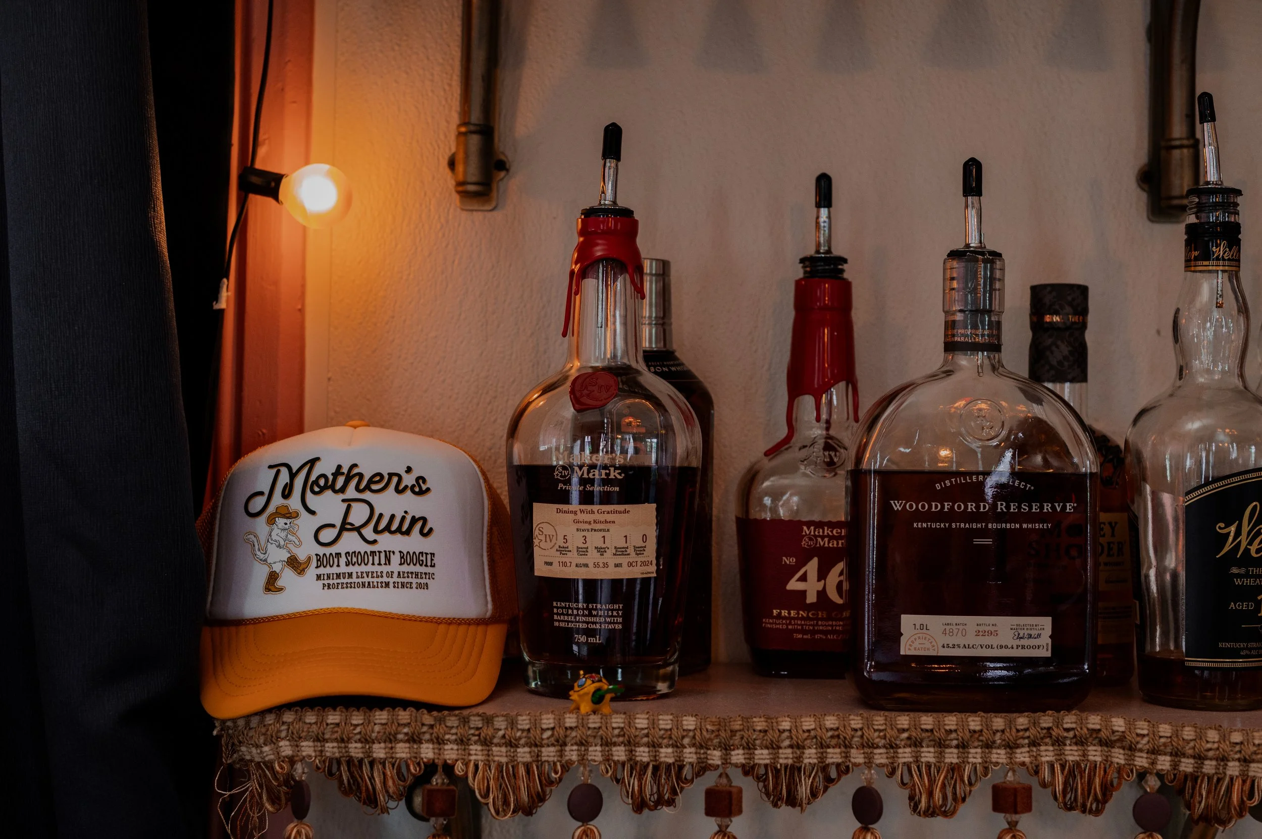 A row of liquor bottles on a shelf with a string of lights and a decorative hat or cap beneath them.