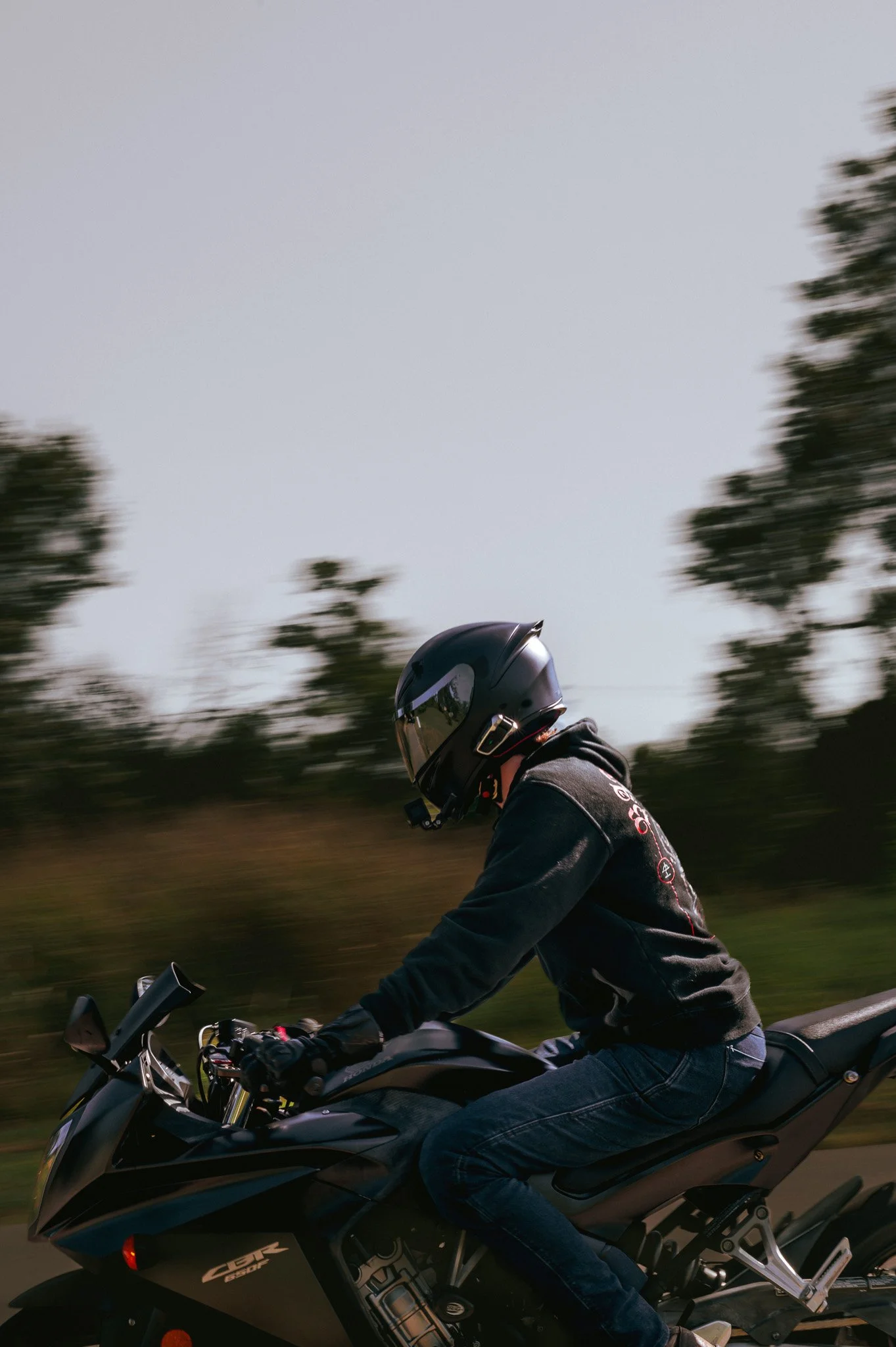 A person riding a black motorcycle on a road with trees in the background and wearing a black helmet and a black jacket.