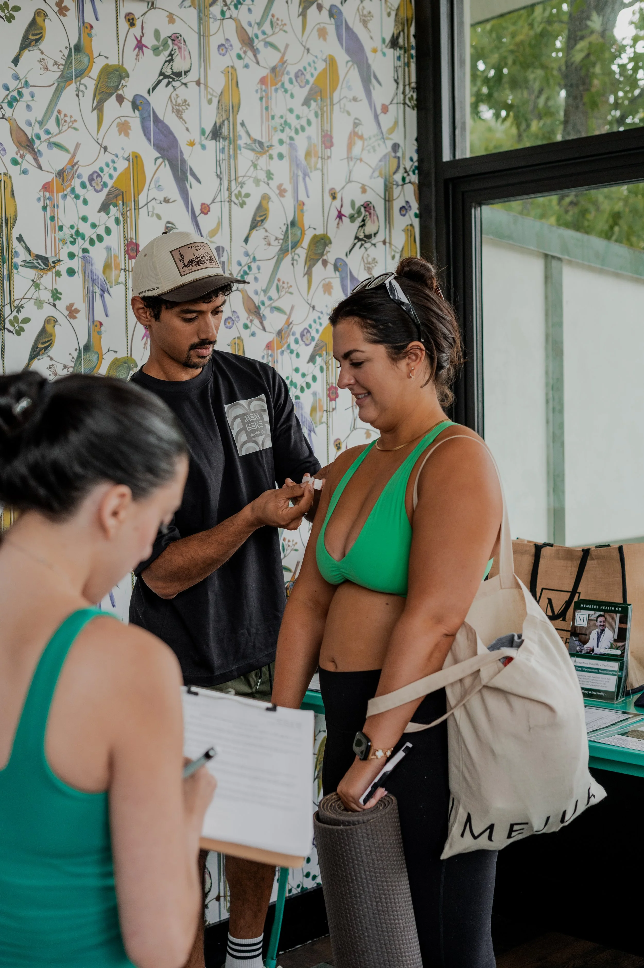 A woman in a green sports bra and black pants checks in at a health clinic while a man assists her. They are inside a room with a colorful bird-themed wallpaper and a large window showing trees outside.