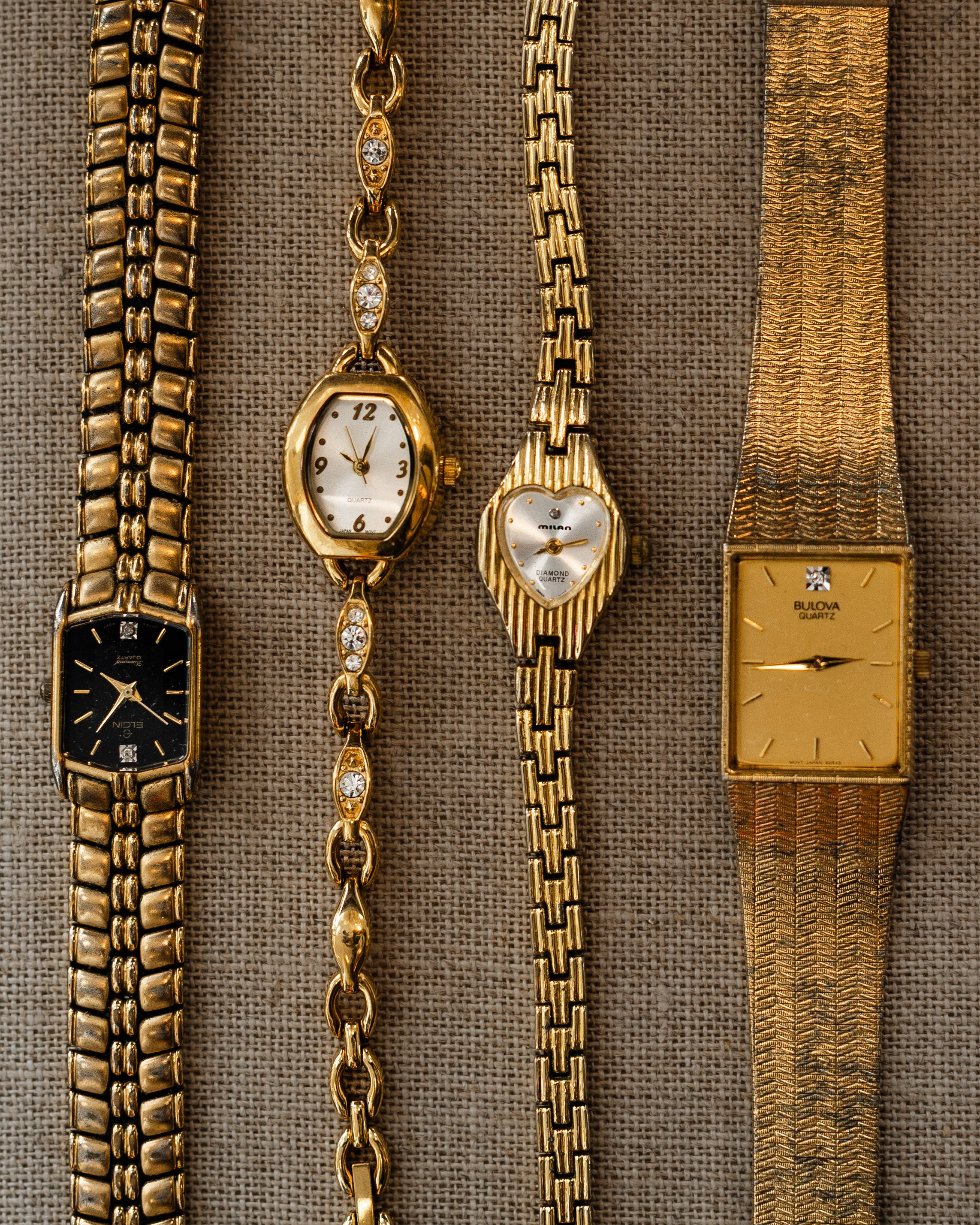 Close-up of five gold watches and bracelets displayed on a textured beige fabric background, showcasing different designs and styles.