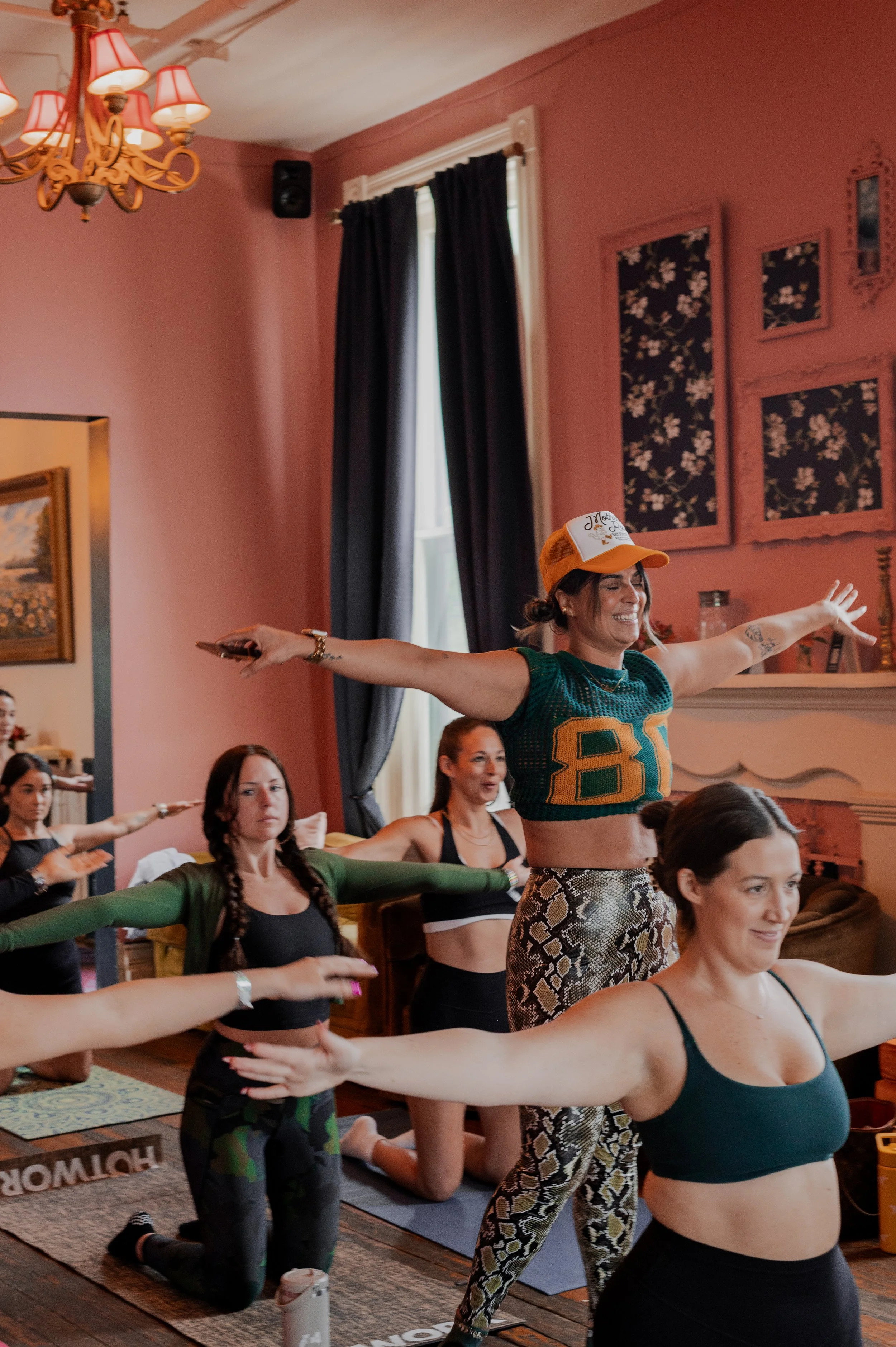 Women participating in a yoga or fitness class in a cozy, vintage-style room with pink walls, floral framed artwork, and a chandelier. Some women are kneeling with arms extended, practicing poses.