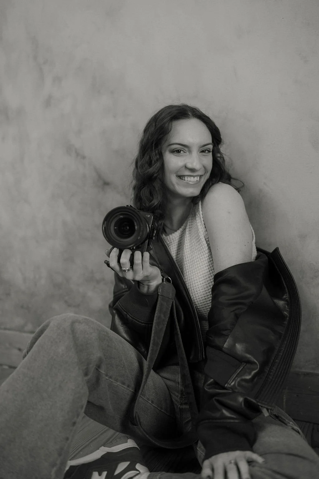 A woman with curly hair smiling while holding a camera, sitting on the floor against a plain wall. She is wearing a sleeveless top and a jacket, with a backpack nearby.