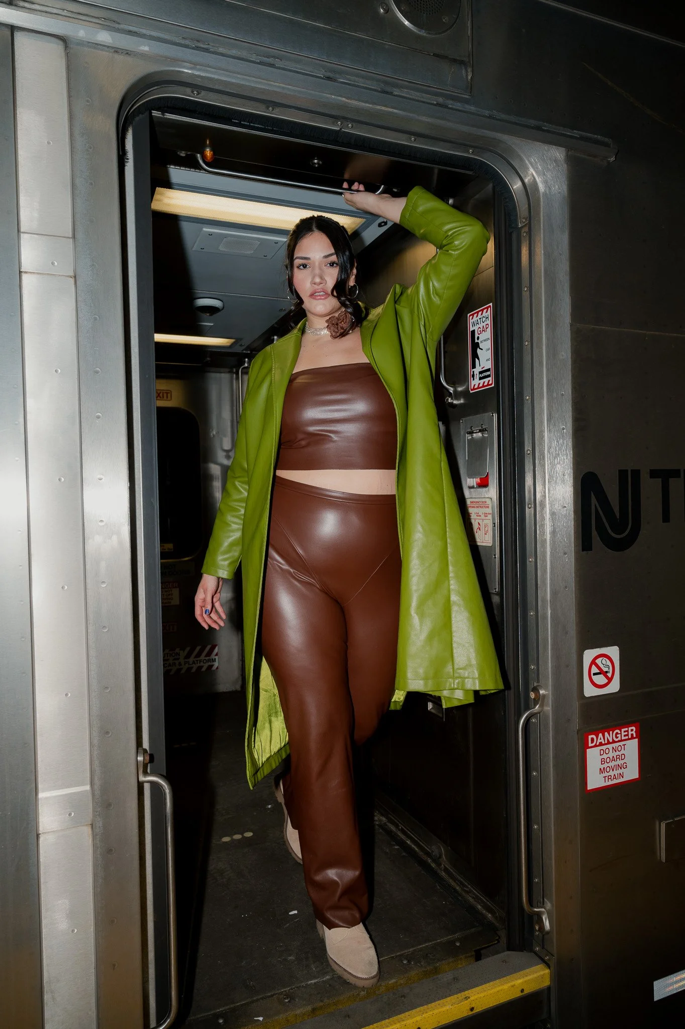 A woman is standing inside a train doorway, slightly leaning out. She is wearing a green coat, brown leather-like top and pants, and beige shoes. She has dark hair and is looking at the camera with a slightly open mouth.