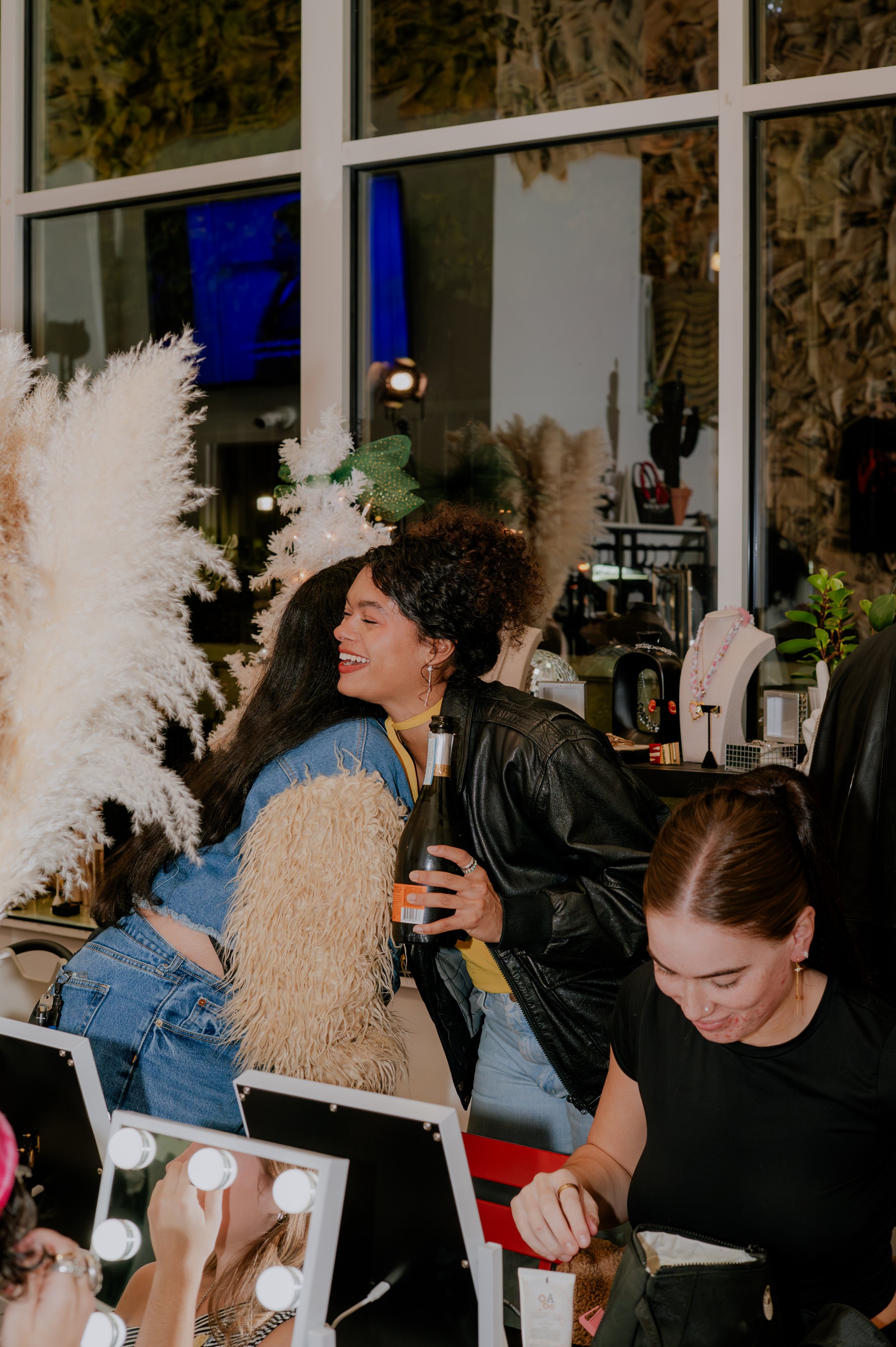 People enjoying themselves at a festive event, with some laughing and engaging in activities at a beauty or jewelry station, decorated with white pampas grass and holiday decorations.