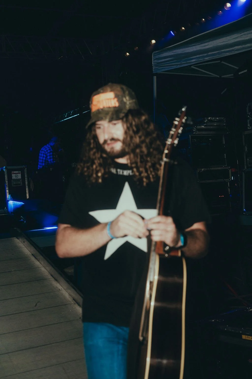 A man with long curly hair, beard, wearing a camouflage baseball cap, black t-shirt with a large white star, and jeans, holding an acoustic guitar at a concert or music event with stage lighting and equipment in the background.
