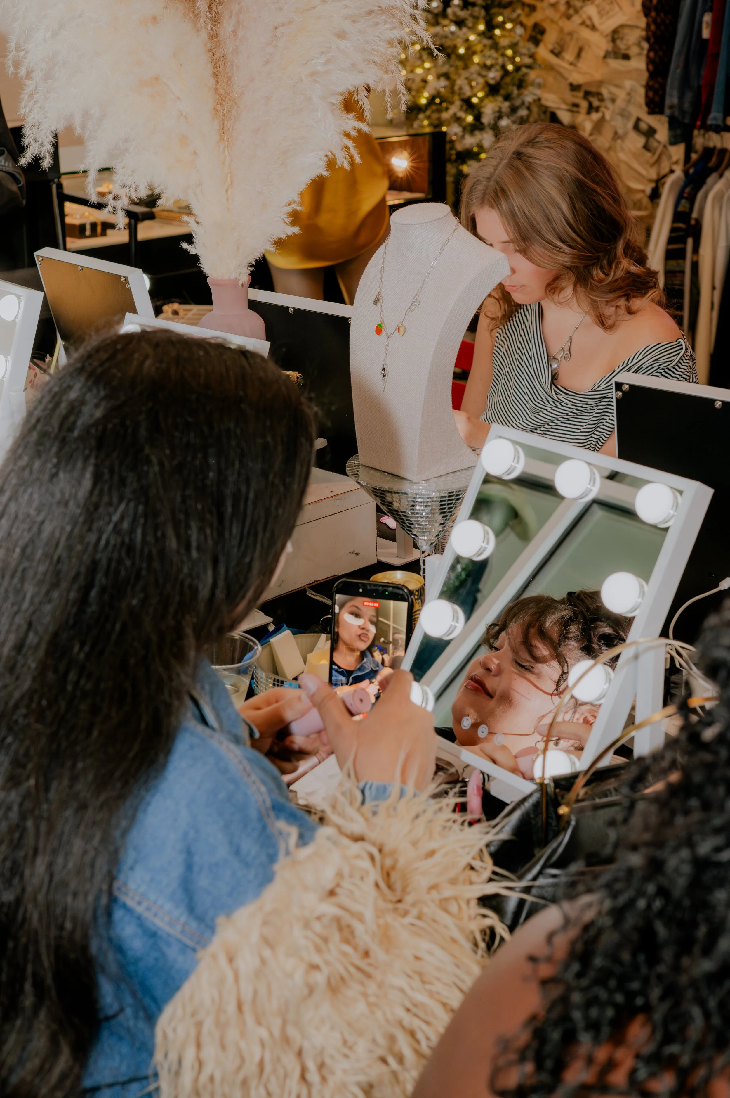 A woman getting foundation applied in front of a mirror with bright Hollywood-style lights, surrounded by vanity makeup supplies, in a beauty salon.