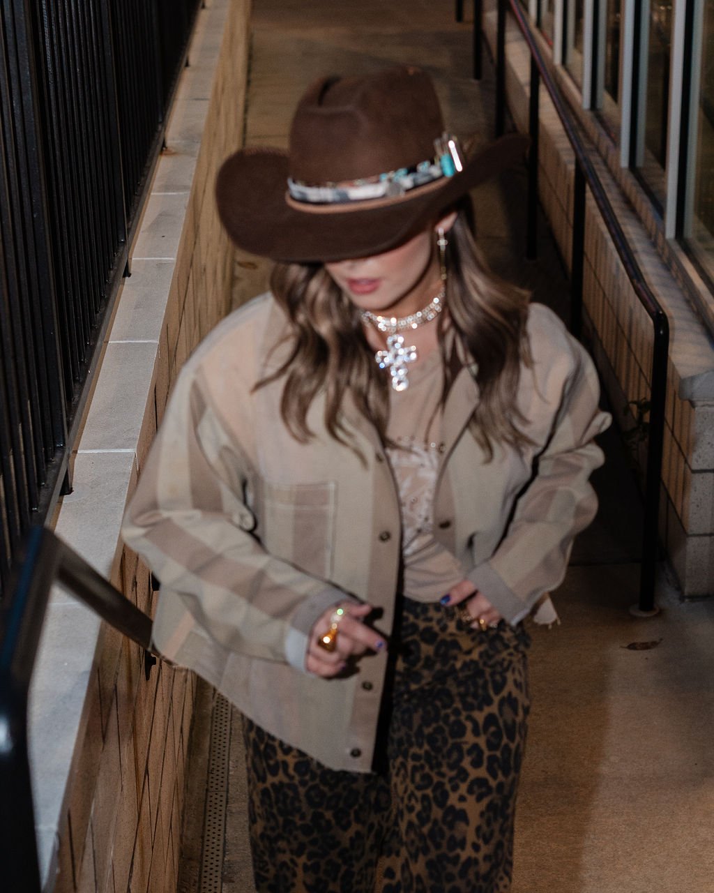 A woman wearing a cowboy hat, beige plaid jacket, leopard print pants, and jewelry, standing indoors next to a brick wall and metal railing.
