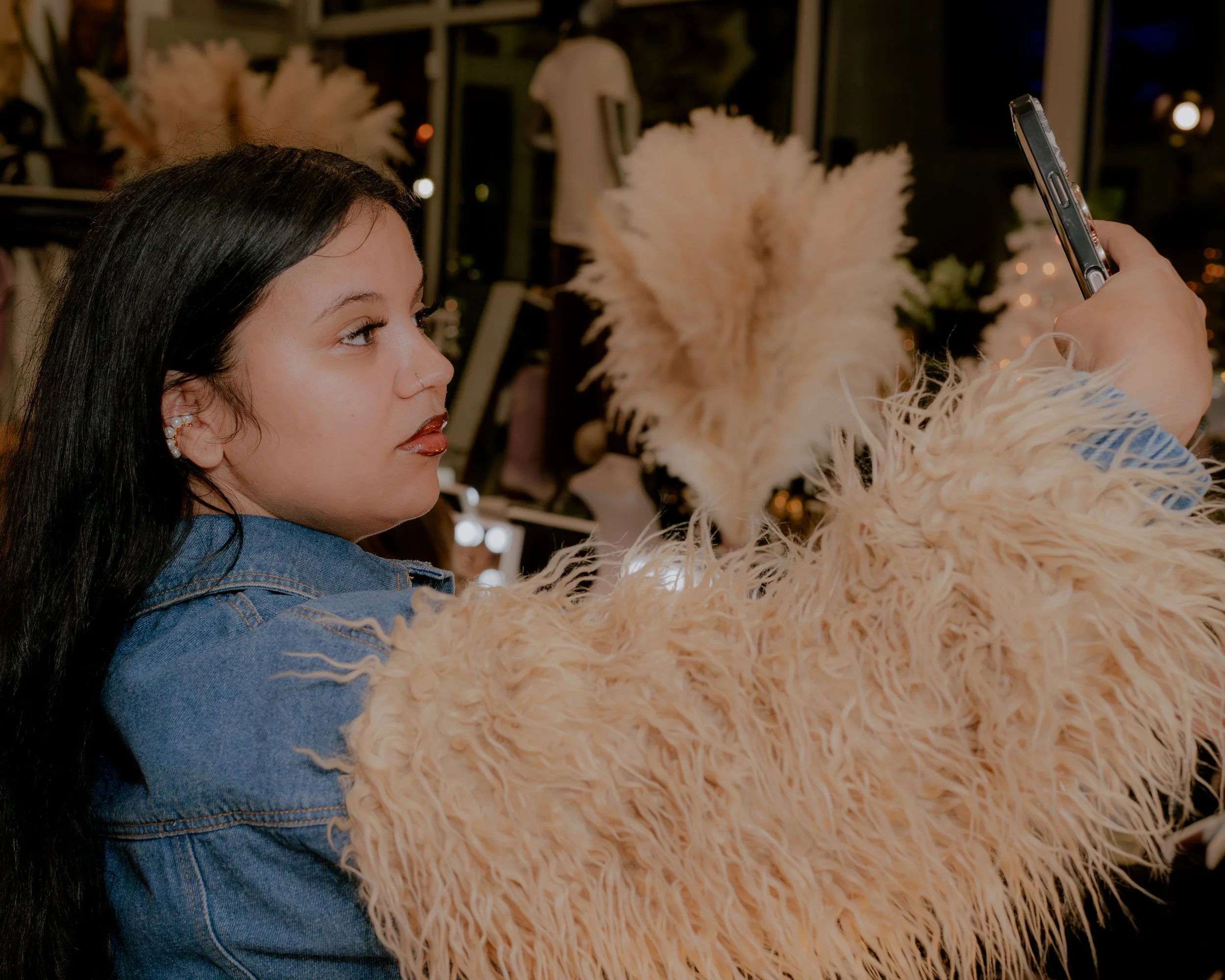 A woman with dark hair and a nose ring is taking a selfie with a person wearing a fluffy beige coat. The background features shelves and decorative items, with warm ambient lighting.