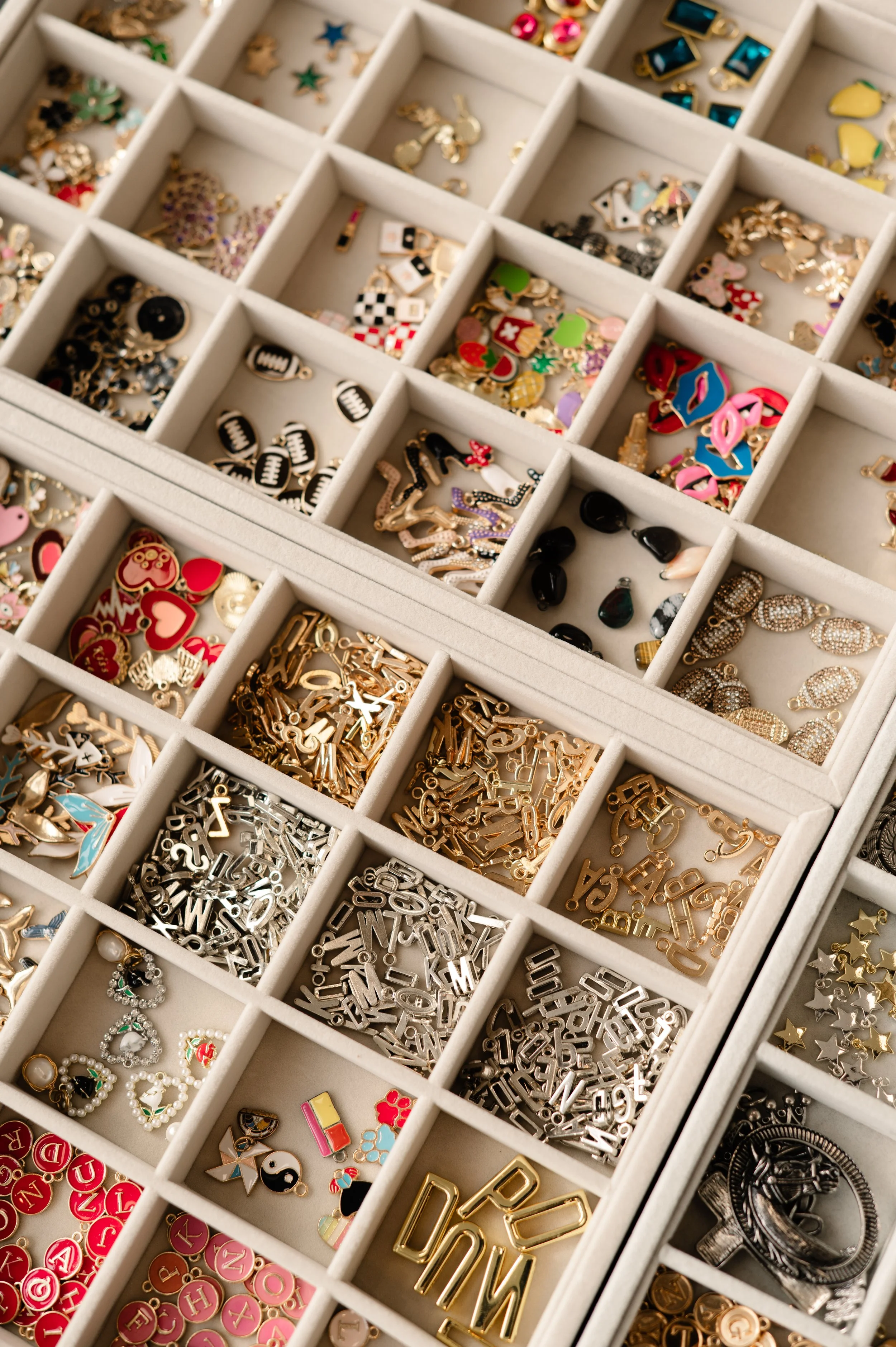 Organized jewelry collection in a compartmentalized display case. Contains various decorative charms, letter charms, themed pendants, and jewelry pieces in gold, silver, and colorful designs.