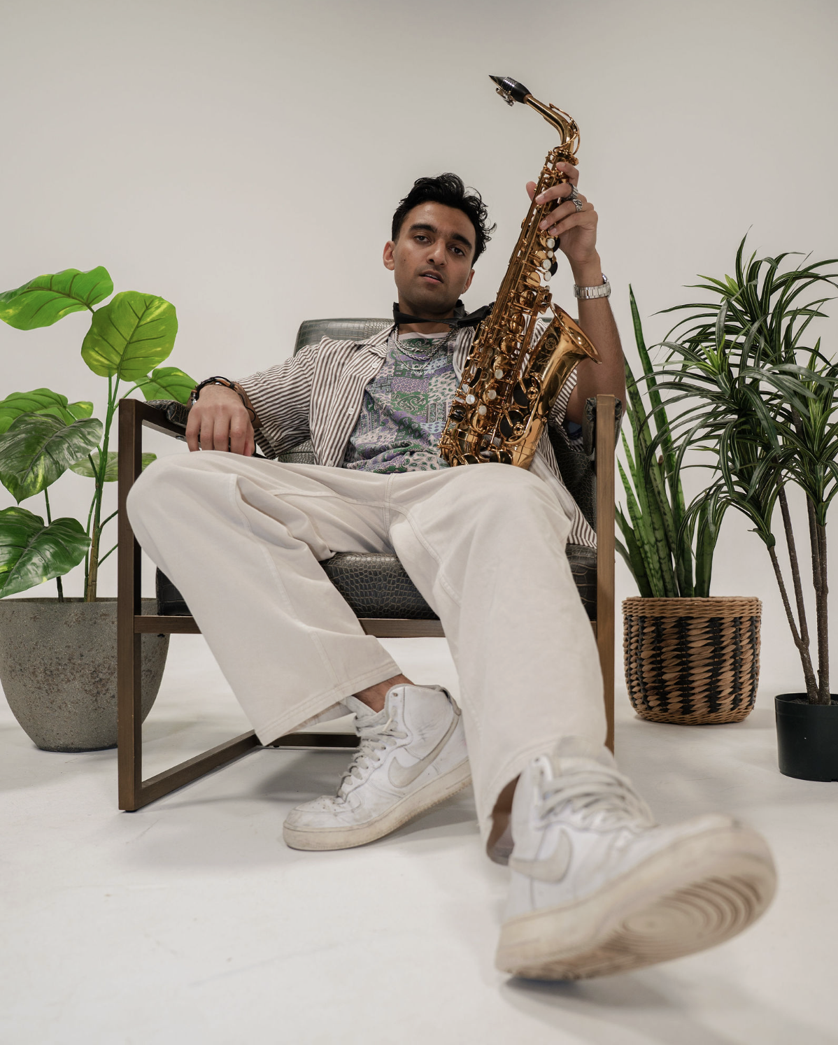 Young man sitting in a chair holding a gold saxophone, wearing white sneakers, light-colored pants, a patterned shirt, and a striped open shirt, surrounded by potted plants in a minimalistic setting.