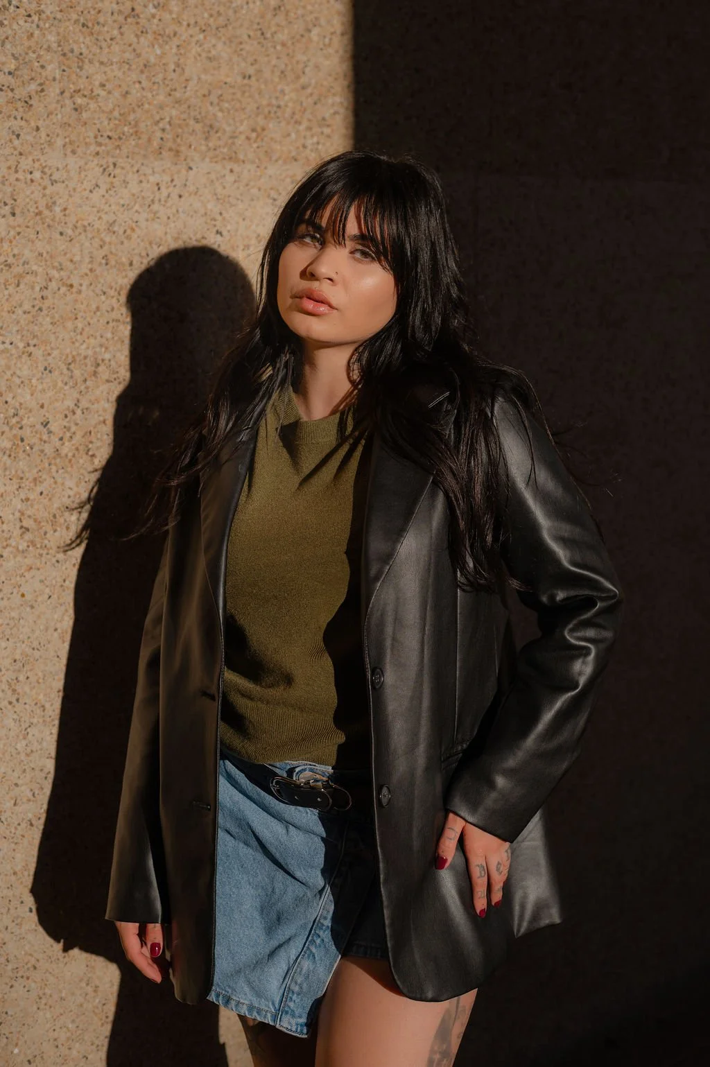 A young woman with long black hair and bangs standing against a textured beige and black wall. She is wearing a black leather jacket over an olive green top, with a denim skirt and a belt. She has tattoos on her hand and red nail polish, and is looking confidently at the camera with a neutral expression.