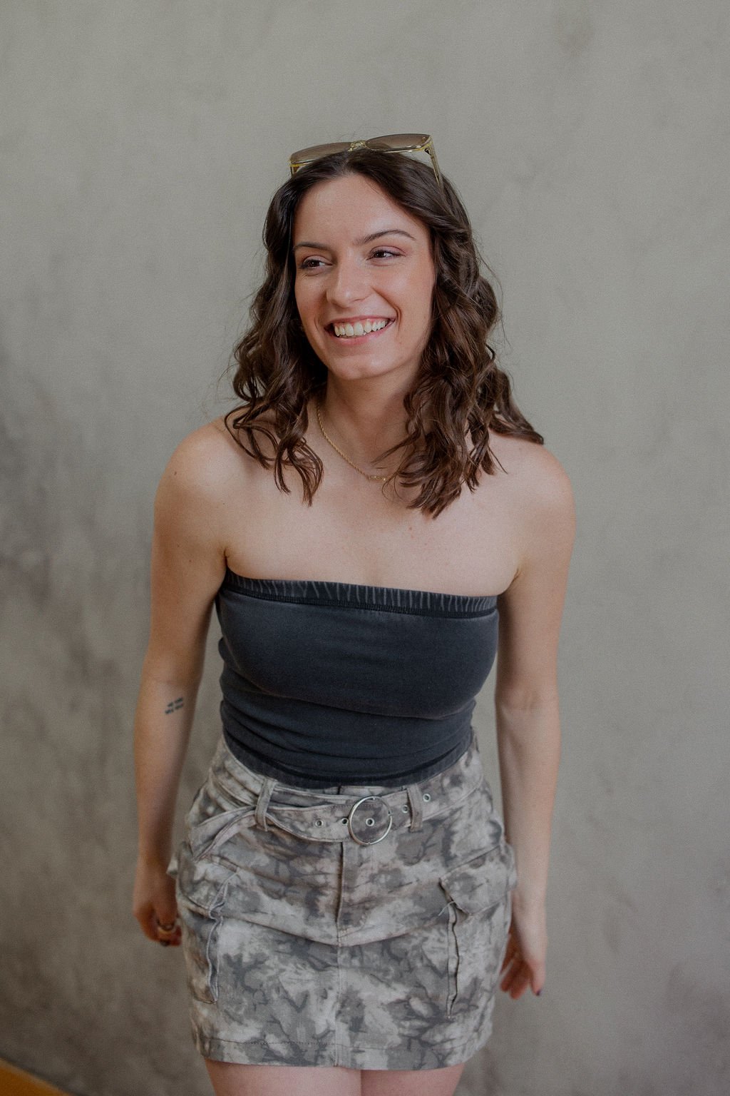 A young woman with shoulder-length curly brown hair, wearing sunglasses on her head, smiling, dressed in a strapless black top and camouflage shorts with a belt, standing against a plain wall.