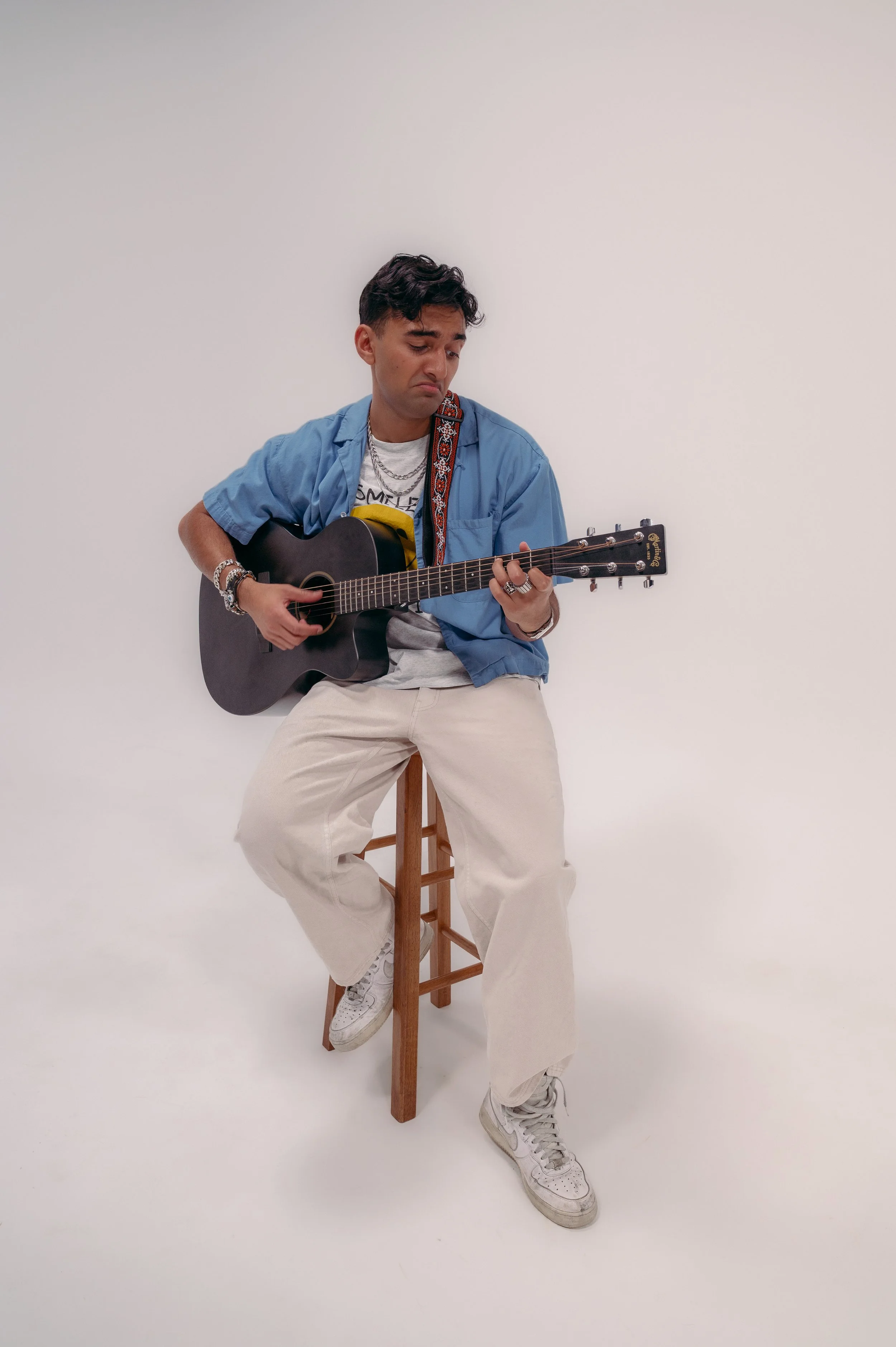 A man sitting on a stool against a plain white background, playing an acoustic guitar. He is wearing a blue short-sleeved shirt over a graphic t-shirt, beige pants, and white sneakers, with accessories such as rings, bracelets, and layered necklaces.