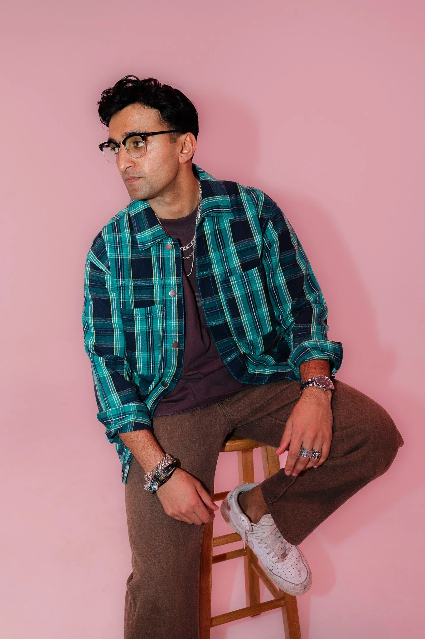 A young man with dark wavy hair and glasses, wearing a green and blue plaid shirt over a dark T-shirt, is sitting on a wooden stool against a pink background. He has accessories including rings and bracelets, and is dressed in brown pants and white s