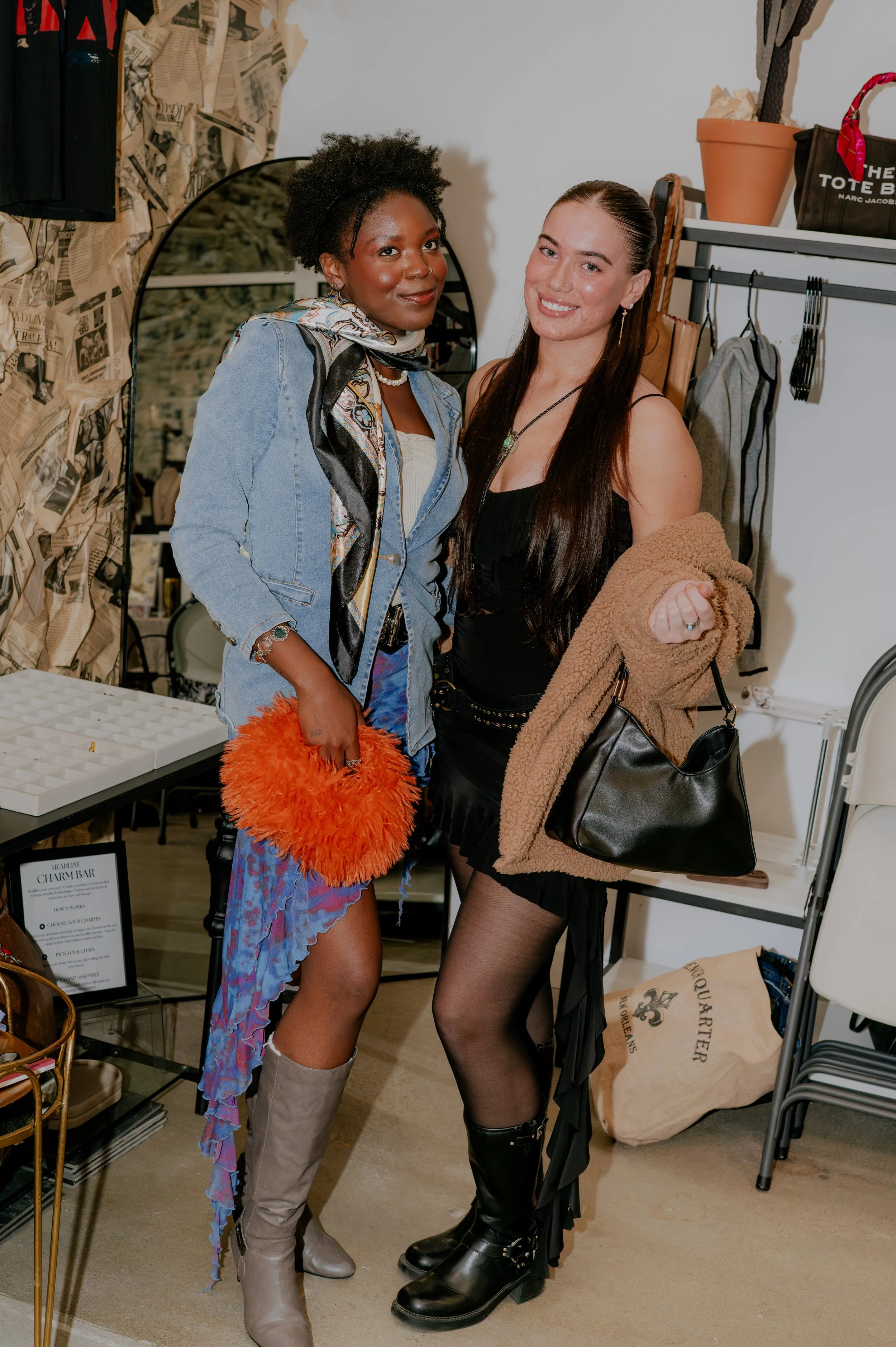 Two women standing indoors near clothing and accessories. One woman has dark skin, curly hair, a denim jacket, and colorful skirt holding an orange fuzzy bag. The other woman has light skin, straight hair, black dress, and black lace-up boots, holdin