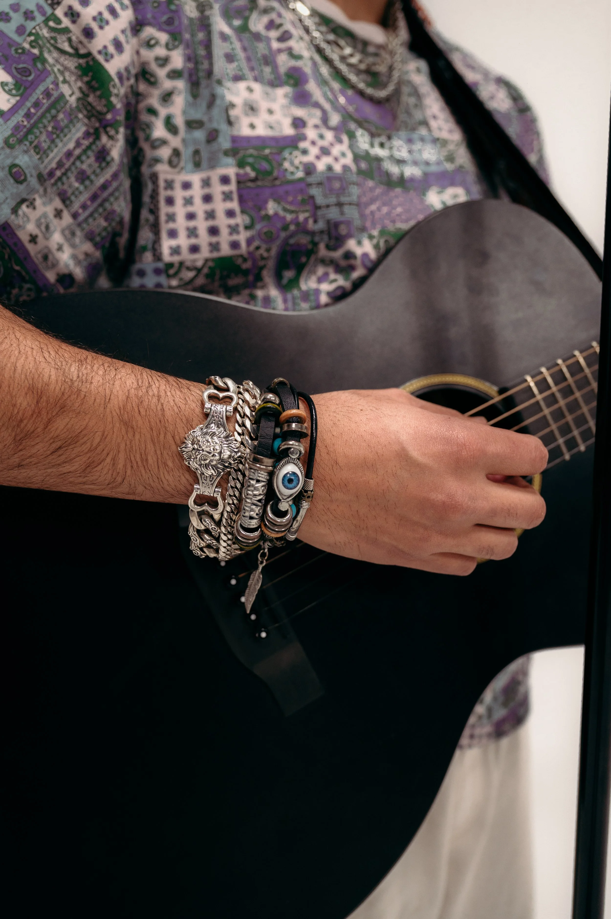 A person playing black acoustic guitar, wearing a colorful patterned shirt and a silver lion wristwatch with layered bracelets with beads, leather, and metal charms.