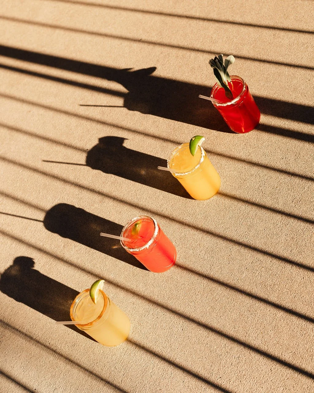 Three colorful cocktails with lime wedges on a beige outdoor surface, casting long shadows from a nearby fence or blinds.