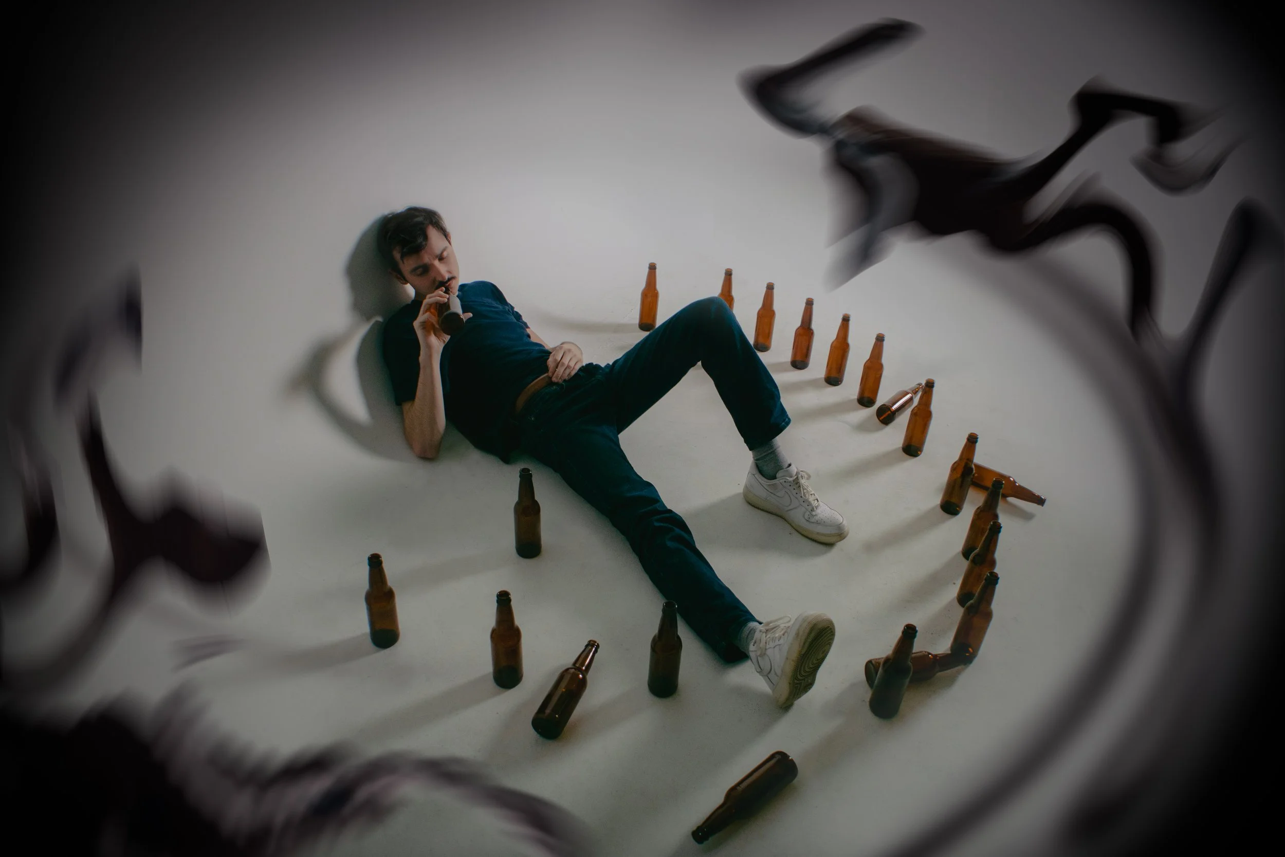 A young man with dark hair lying on the floor among empty brown beer bottles, holding a beer bottle to his mouth, seen through a fractured mirror.