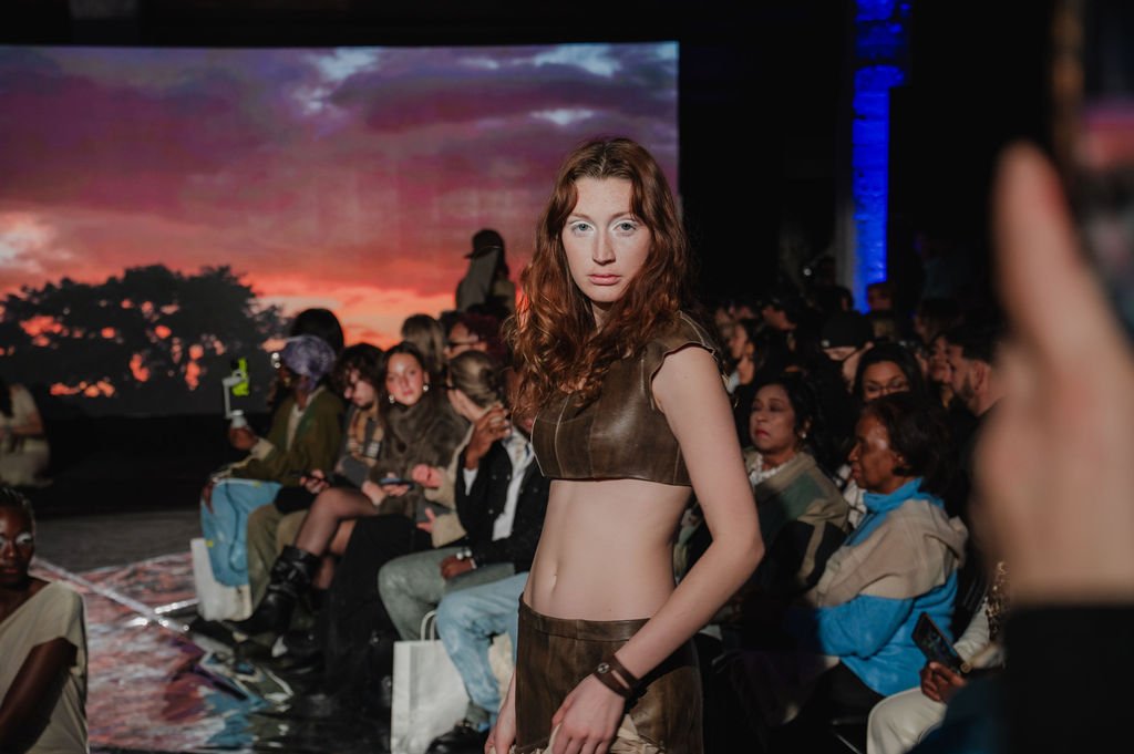 A young woman with long, curly red hair standing in front of an audience during a fashion show at sunset. She wears a brown leather crop top and skirt, with a bracelet on her wrist, and looks directly at the camera. The audience behind her is seated, some engaged with their phones, with a large screen displaying a sunset scene.
