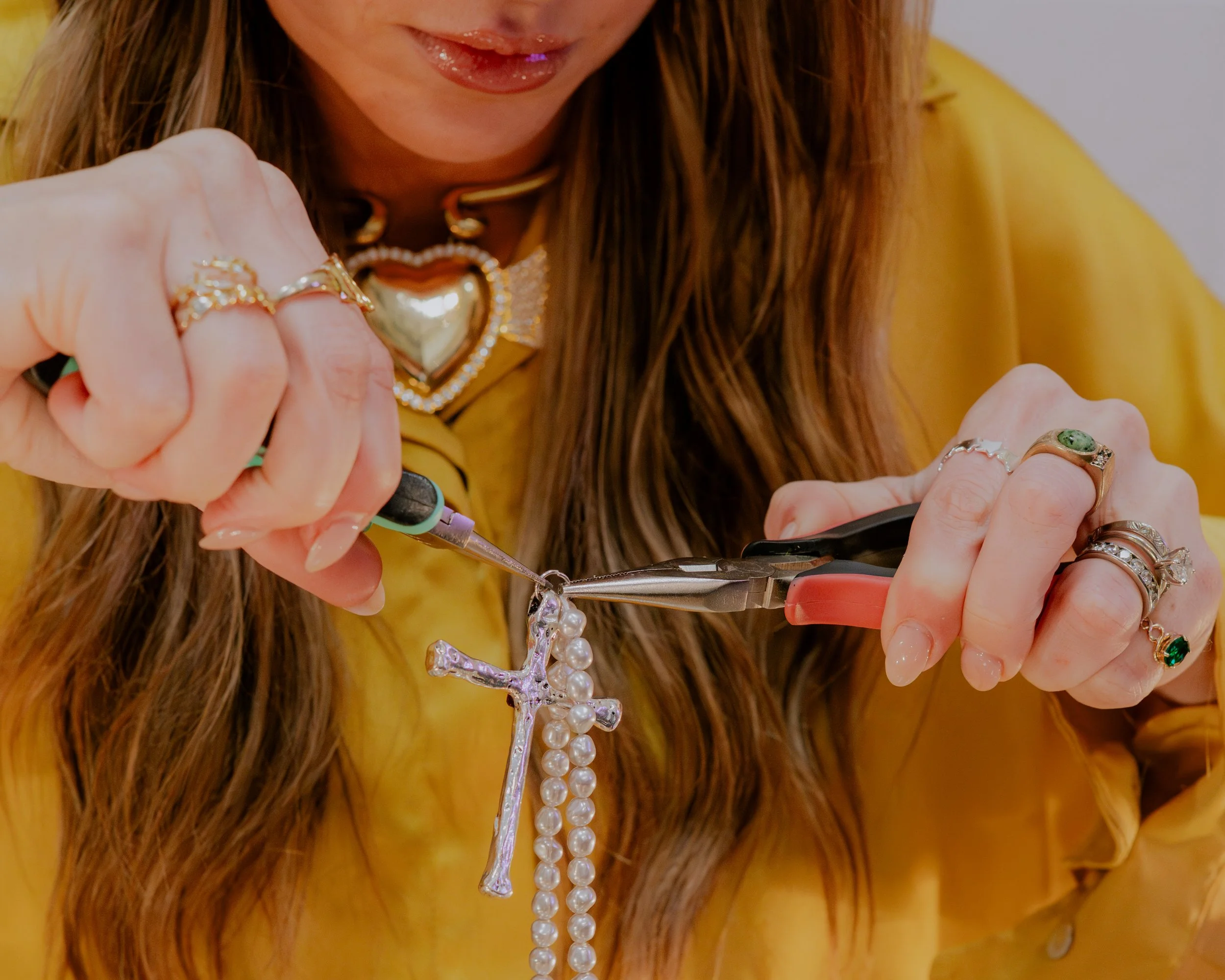 Close-up of a woman in a yellow top attaching a pearl necklace with a fashion cross pendant, wearing multiple rings, including one with a green gemstone, and a large heart-shaped gold necklace.