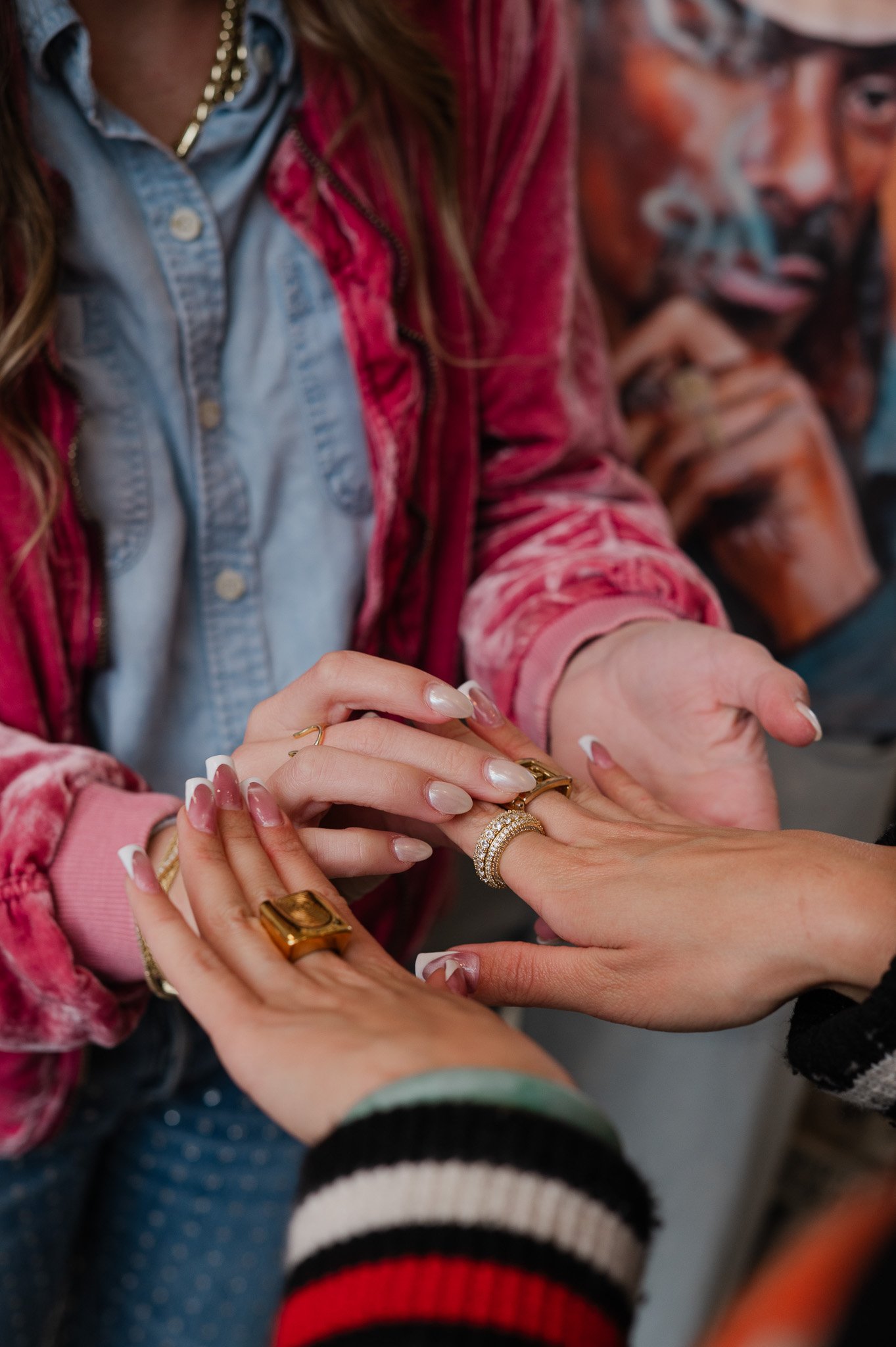 Close-up of multiple women exchanging rings, showing their hands and jewelry, with some wearing rings with diamonds. The women are dressed in colorful, casual clothing, including a denim shirt and pink velvet jacket, and are in an indoor setting.